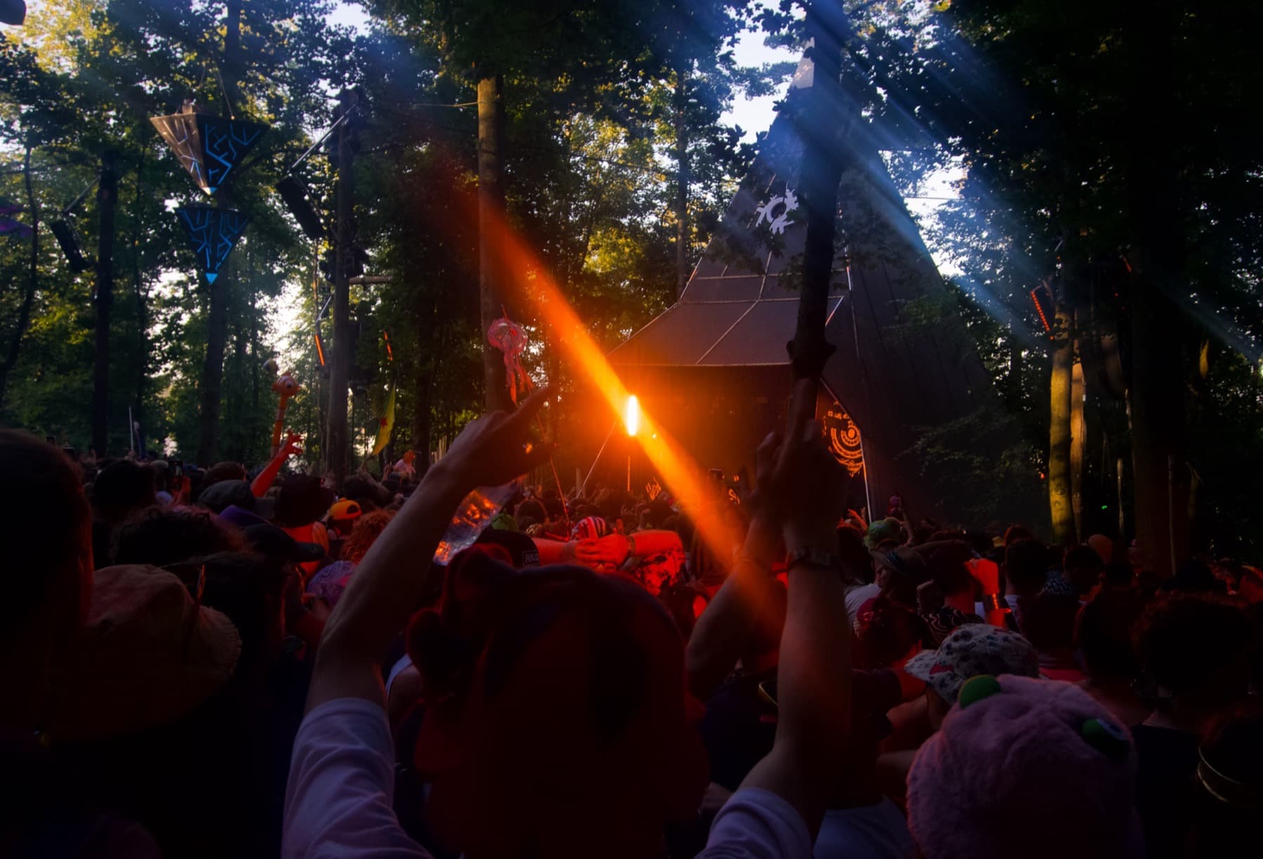 Festival crowd in a woodland clearing with a flare held aloft casting orange light through the trees