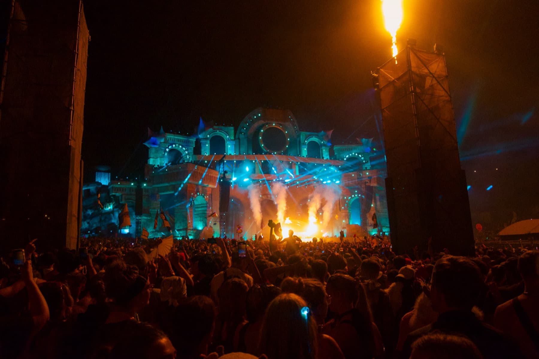Night-time crowd before a grand illuminated stage with teal lights, pyrotechnics, and fire columns