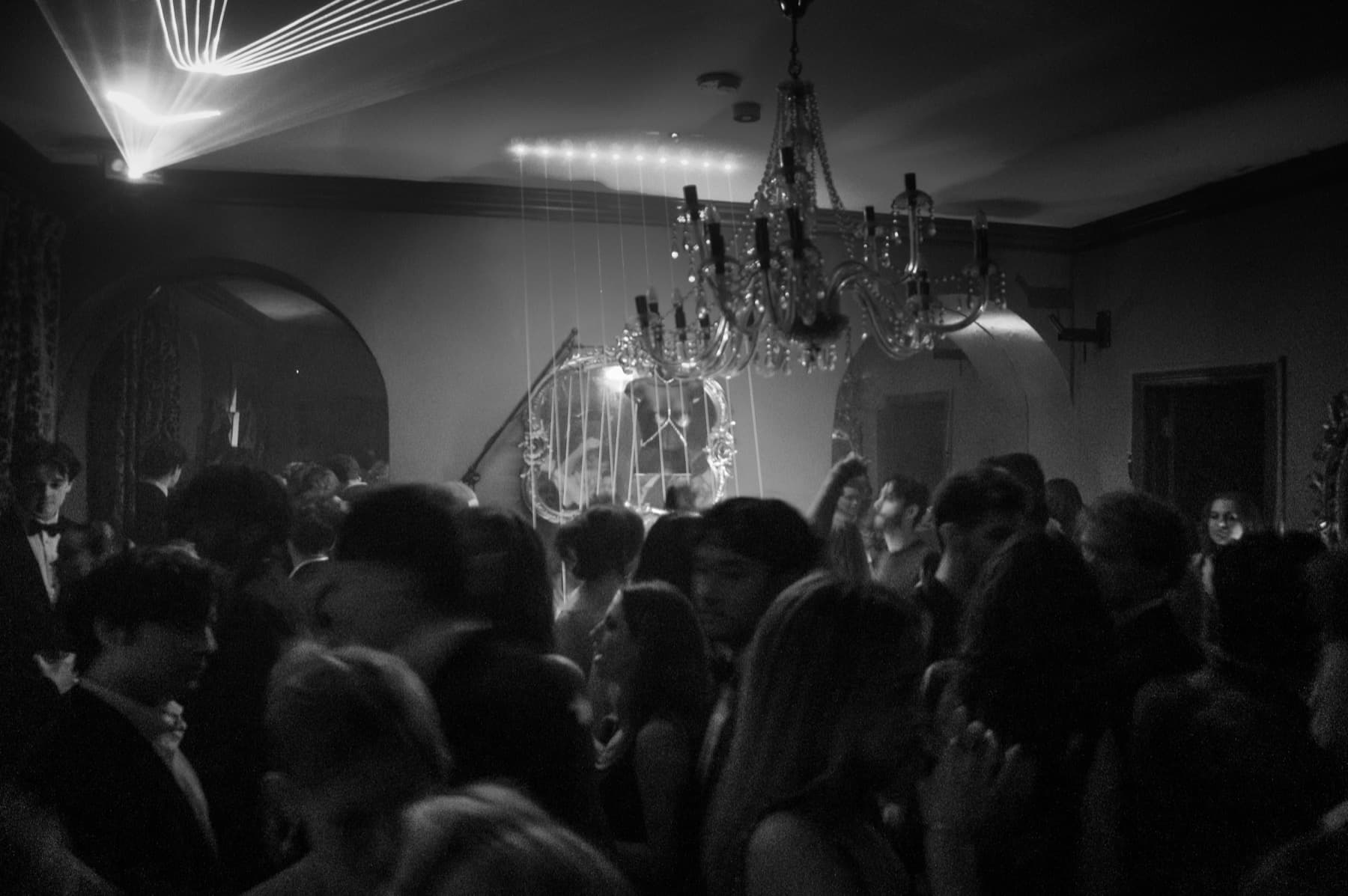 Packed dance floor beneath a crystal chandelier and laser beams in an ornate mansion ballroom