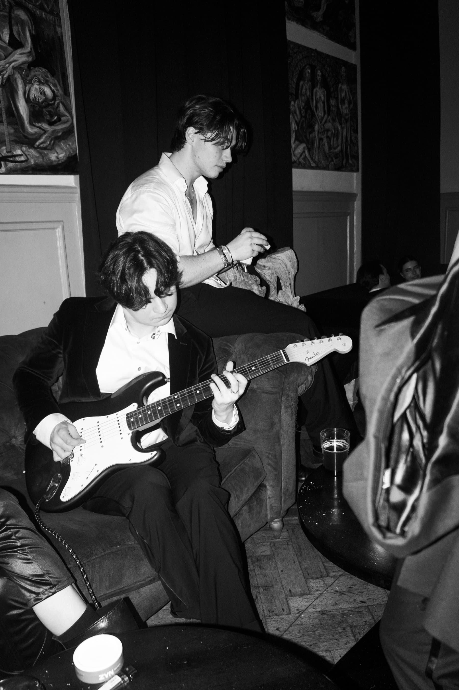 Guitarist playing a Stratocaster on a leather sofa with paintings on the wall behind in black and white