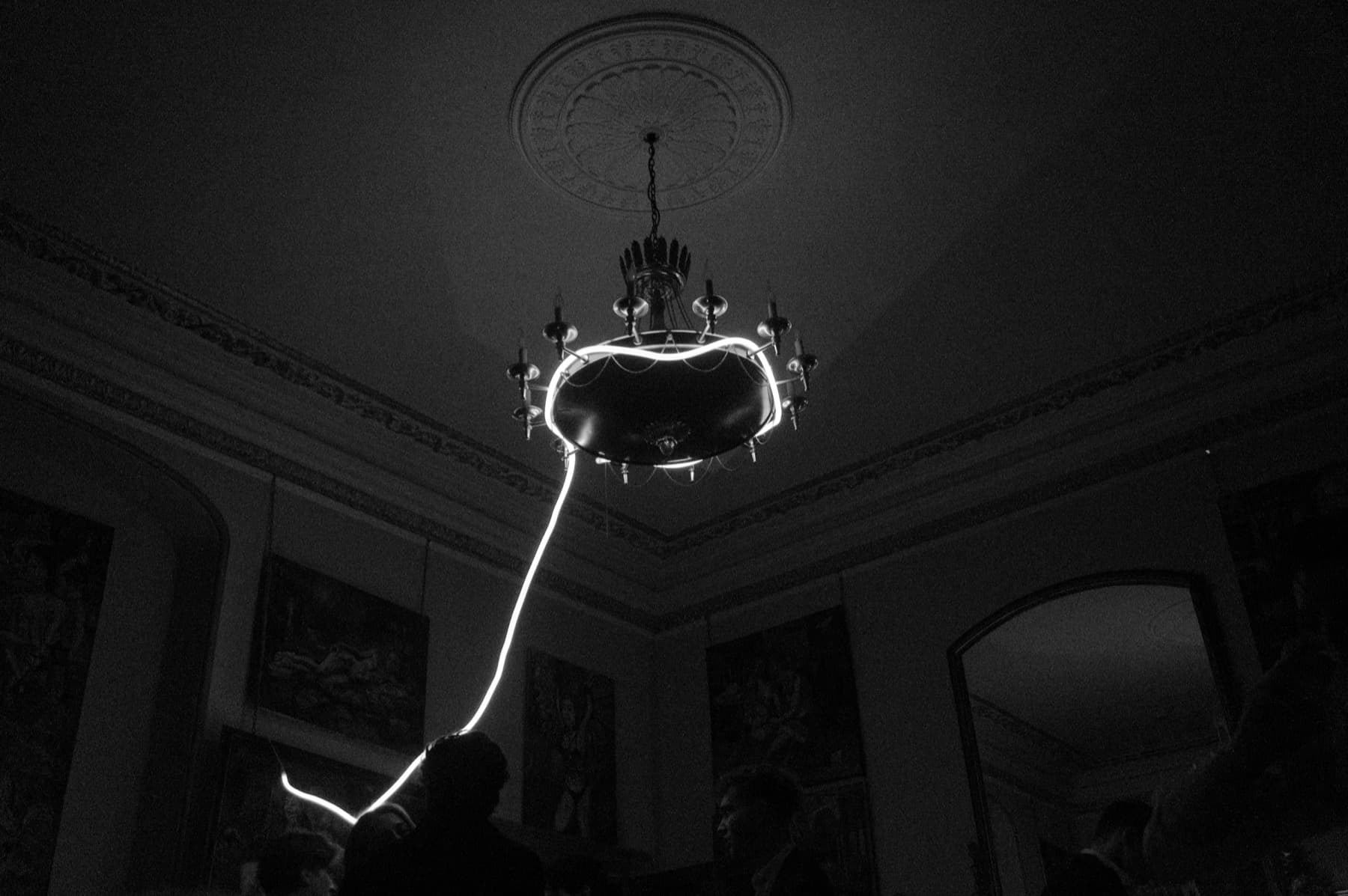 Ornate chandelier on a carved ceiling with a neon light tube trailing down in a grand mansion room