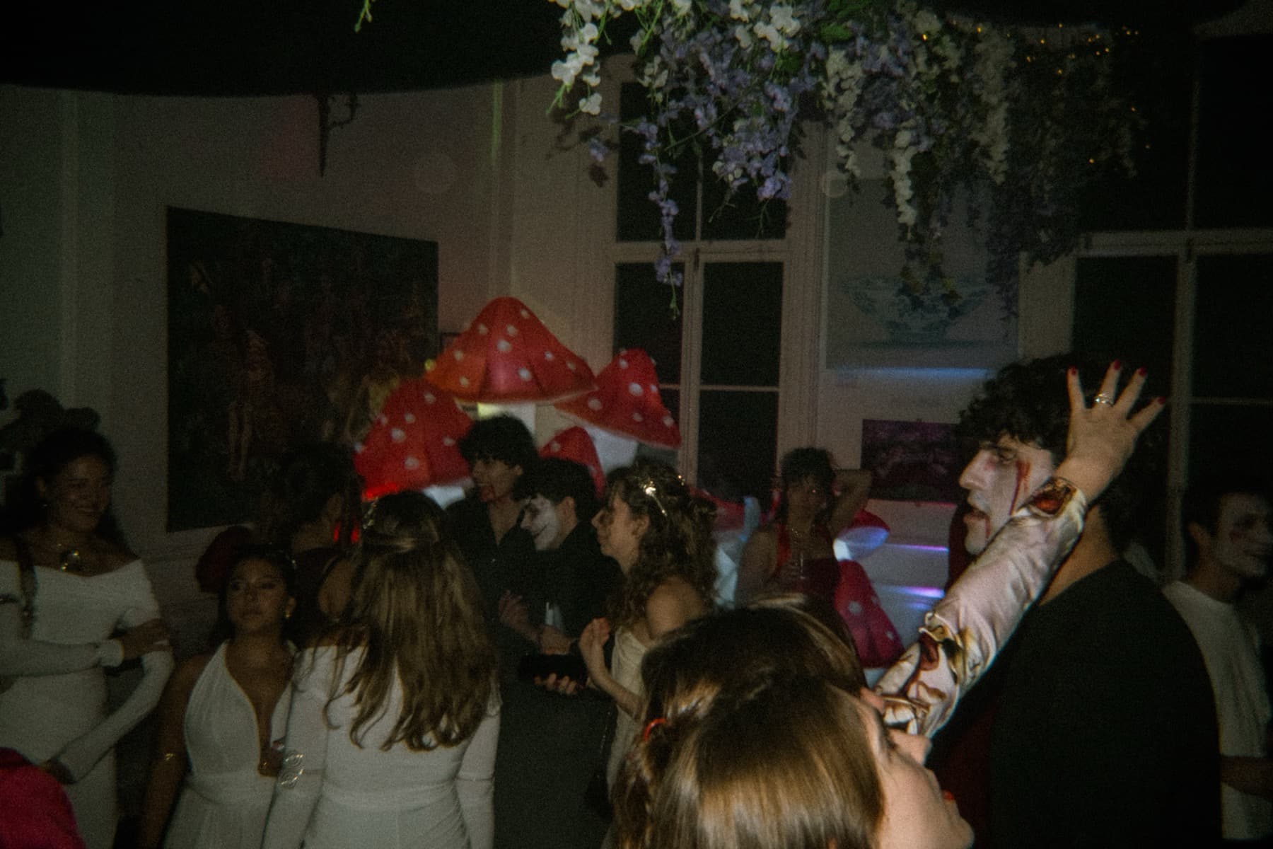 Costumed crowd dancing near giant red-and-white mushroom props and hanging flowers