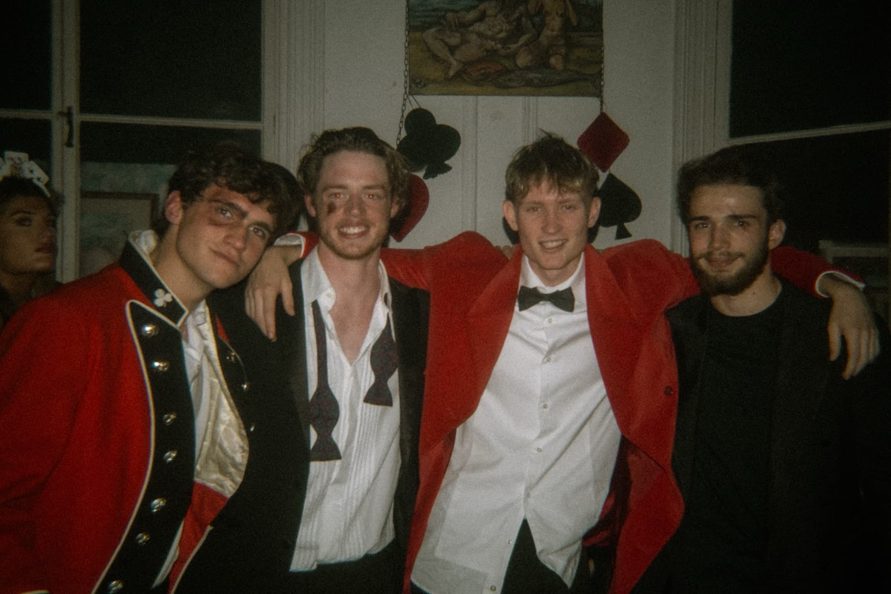 Four friends posing together in red jackets and formal wear with playing card suit decorations on the wall