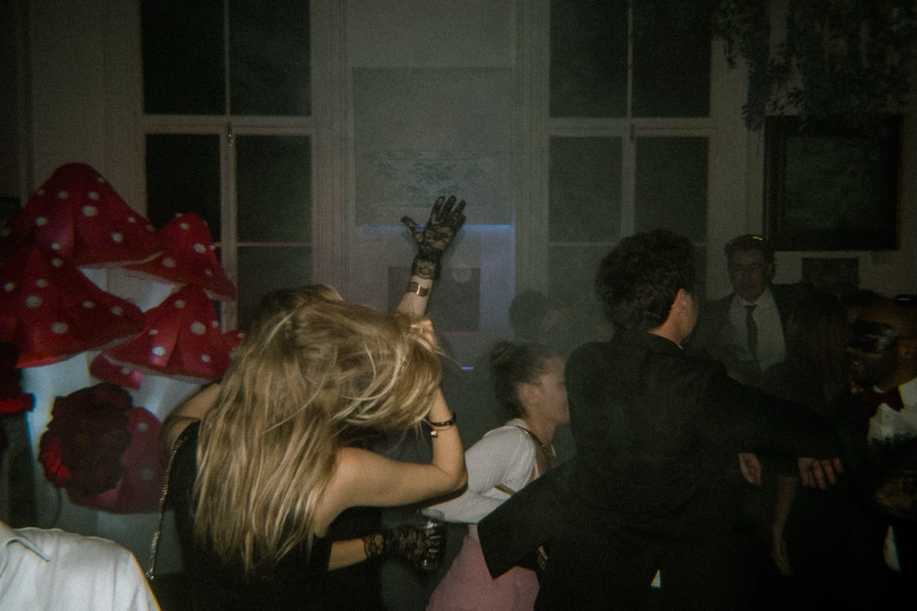 Woman in lace gloves dancing with arm raised beside oversized polka-dot mushroom props in hazy room
