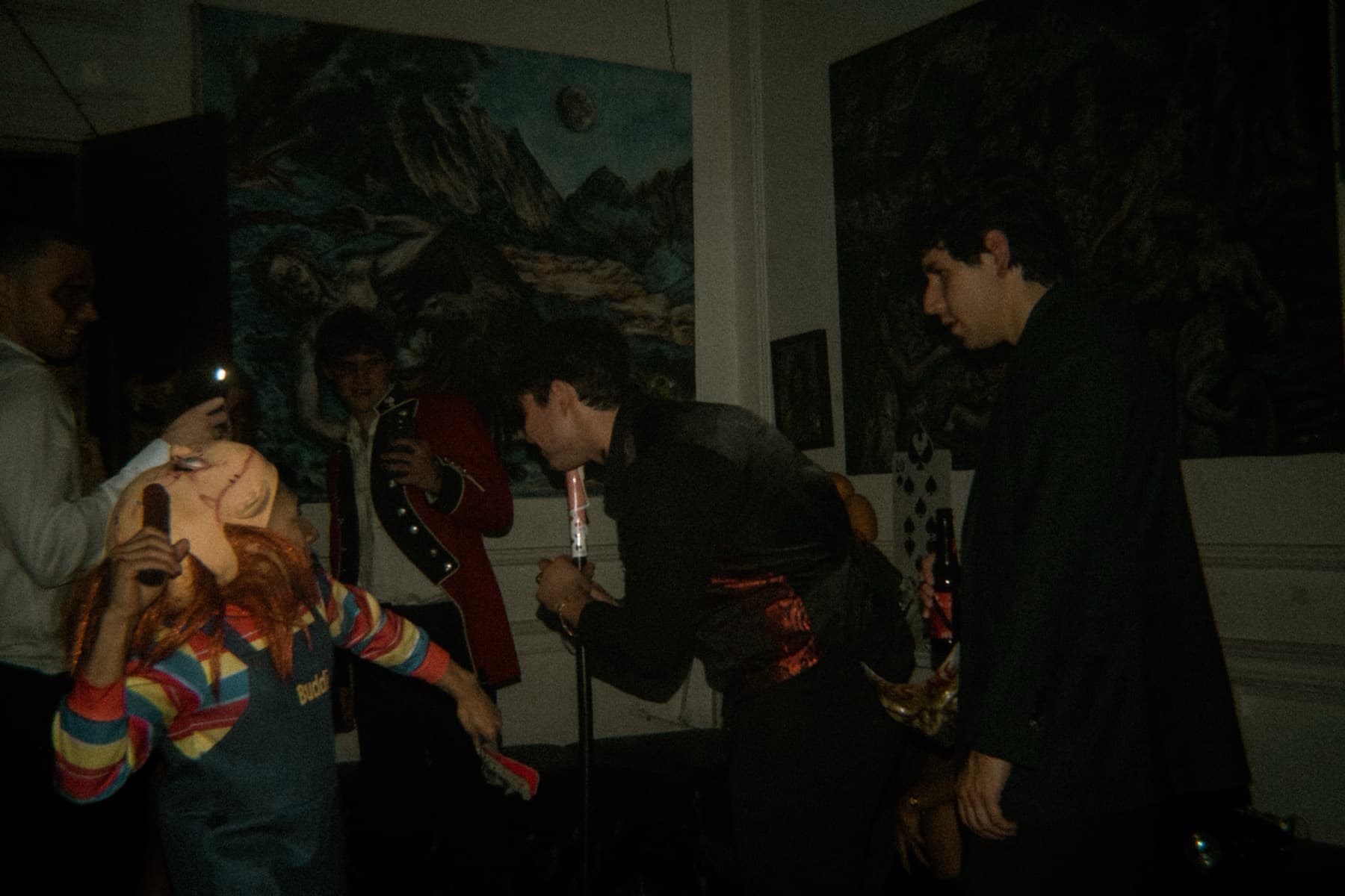 Costumed guests gathered near dark paintings as a Chucky costume wearer interacts with the crowd