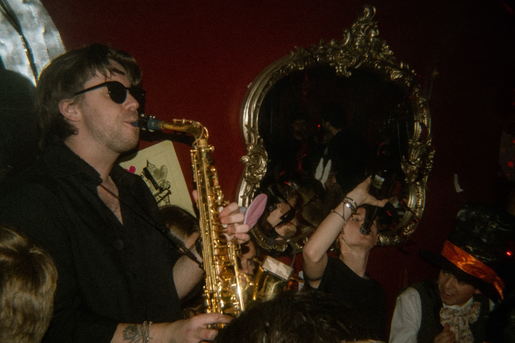 Saxophonist in sunglasses playing among costumed partygoers beside an ornate gold mirror