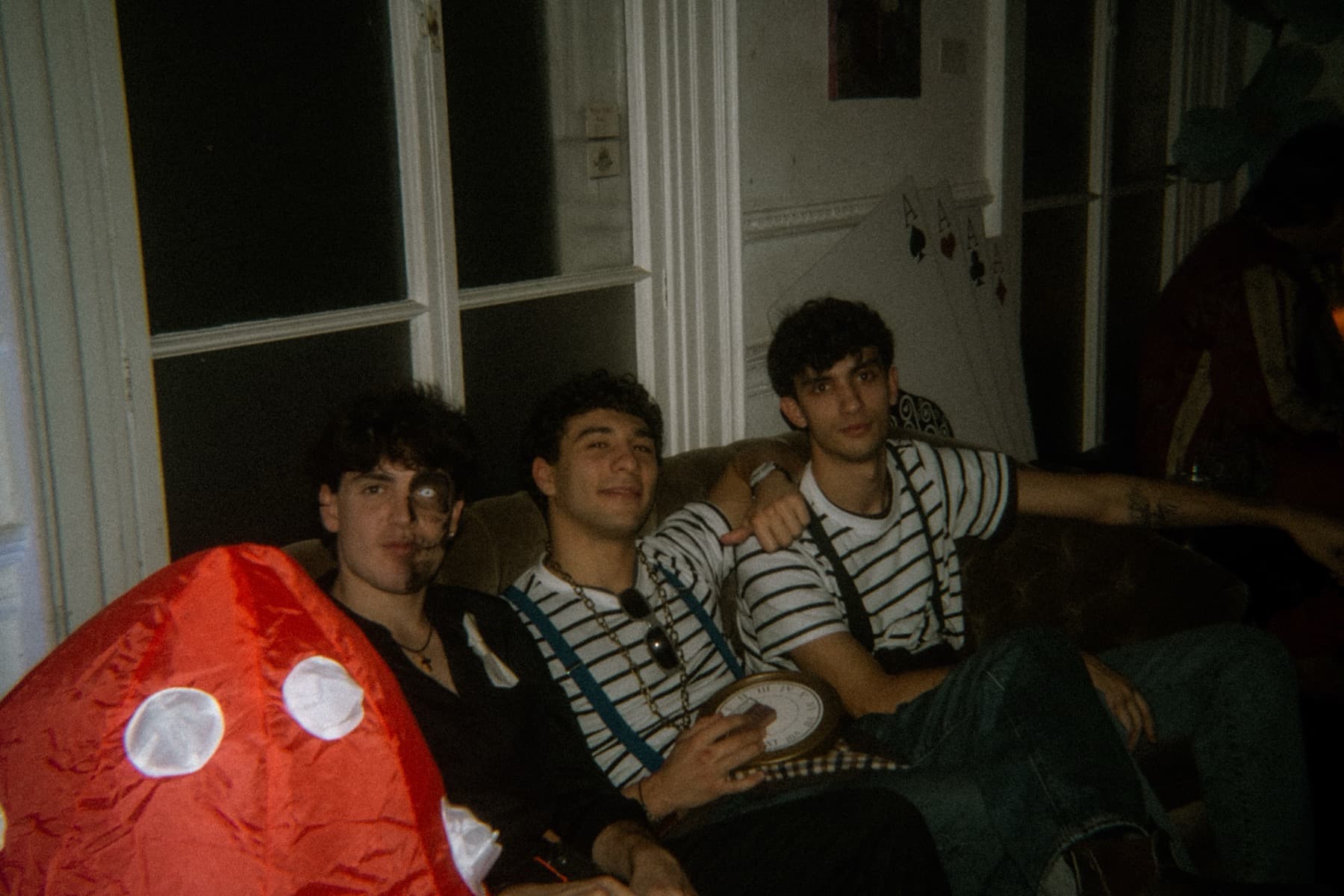Three friends relaxing on a sofa beside a glowing red mushroom prop with striped shirts and face paint