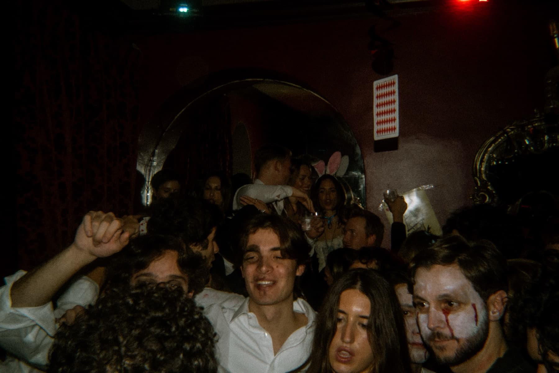 Packed crowd singing along in a red-lit room with an arched mirror and playing card wall art