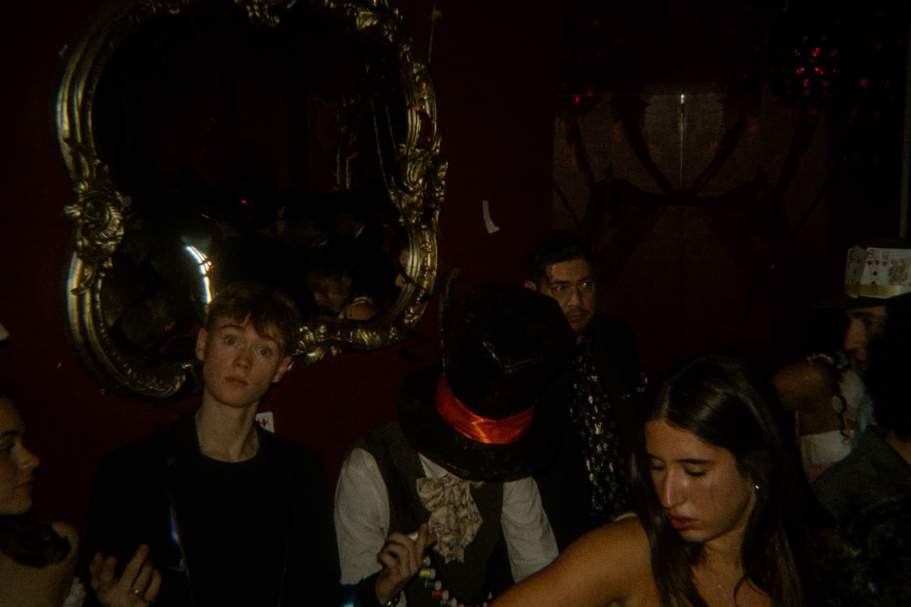 Partygoers in costume near an ornate gold mirror in a deep red room with playing cards on the wall