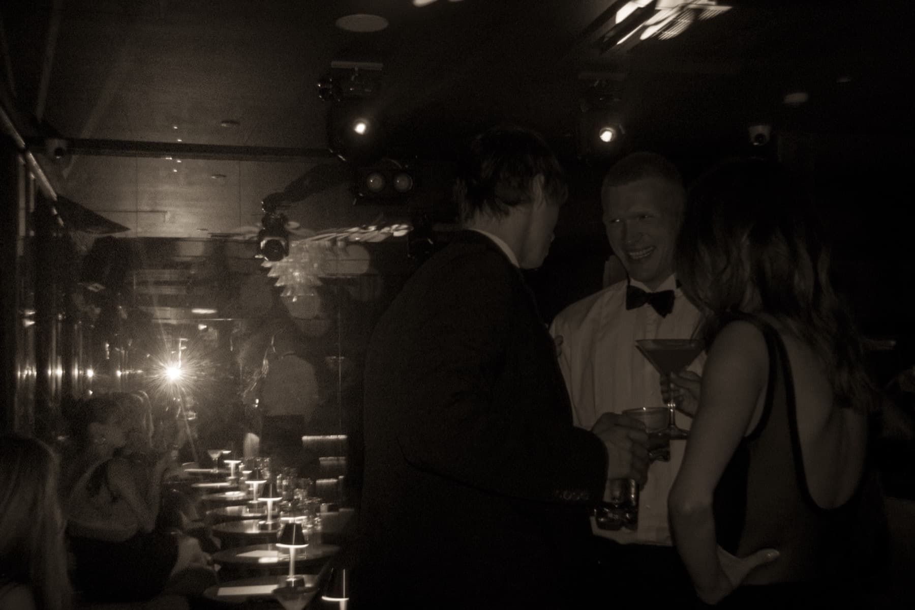 Couple holding a martini glass standing in a moody, dimly lit lounge with a chandelier reflected in the glass wall