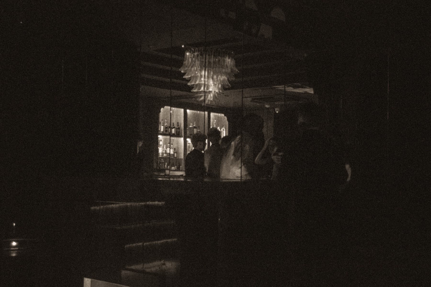 Dimly lit cocktail bar with a glowing chandelier and silhouetted figures at the backlit counter