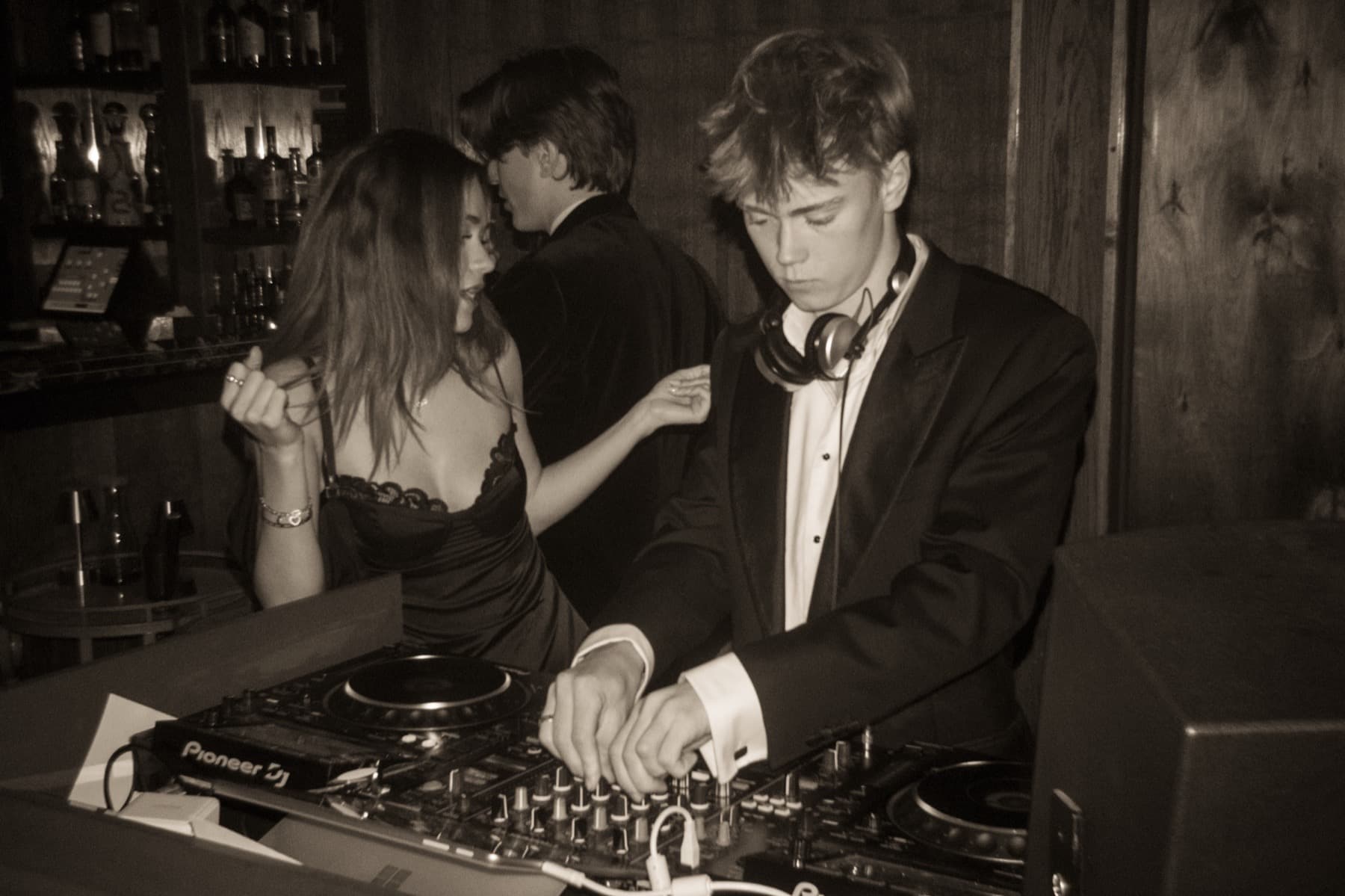 Young DJ in a suit with headphones around his neck focused on Pioneer CDJs as a couple dances behind