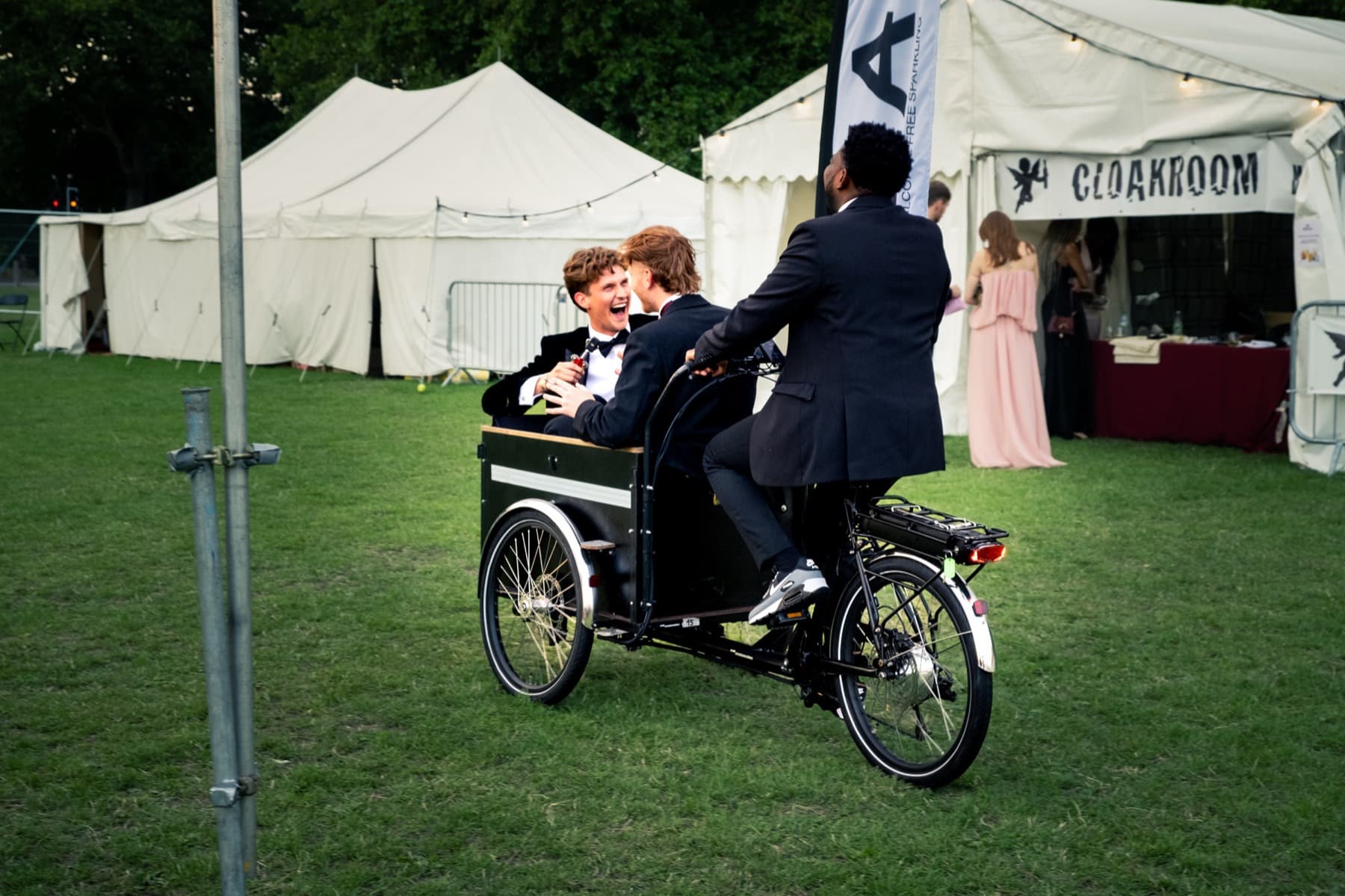 Three friends in black tie laughing while riding a cargo bike across the festival lawn