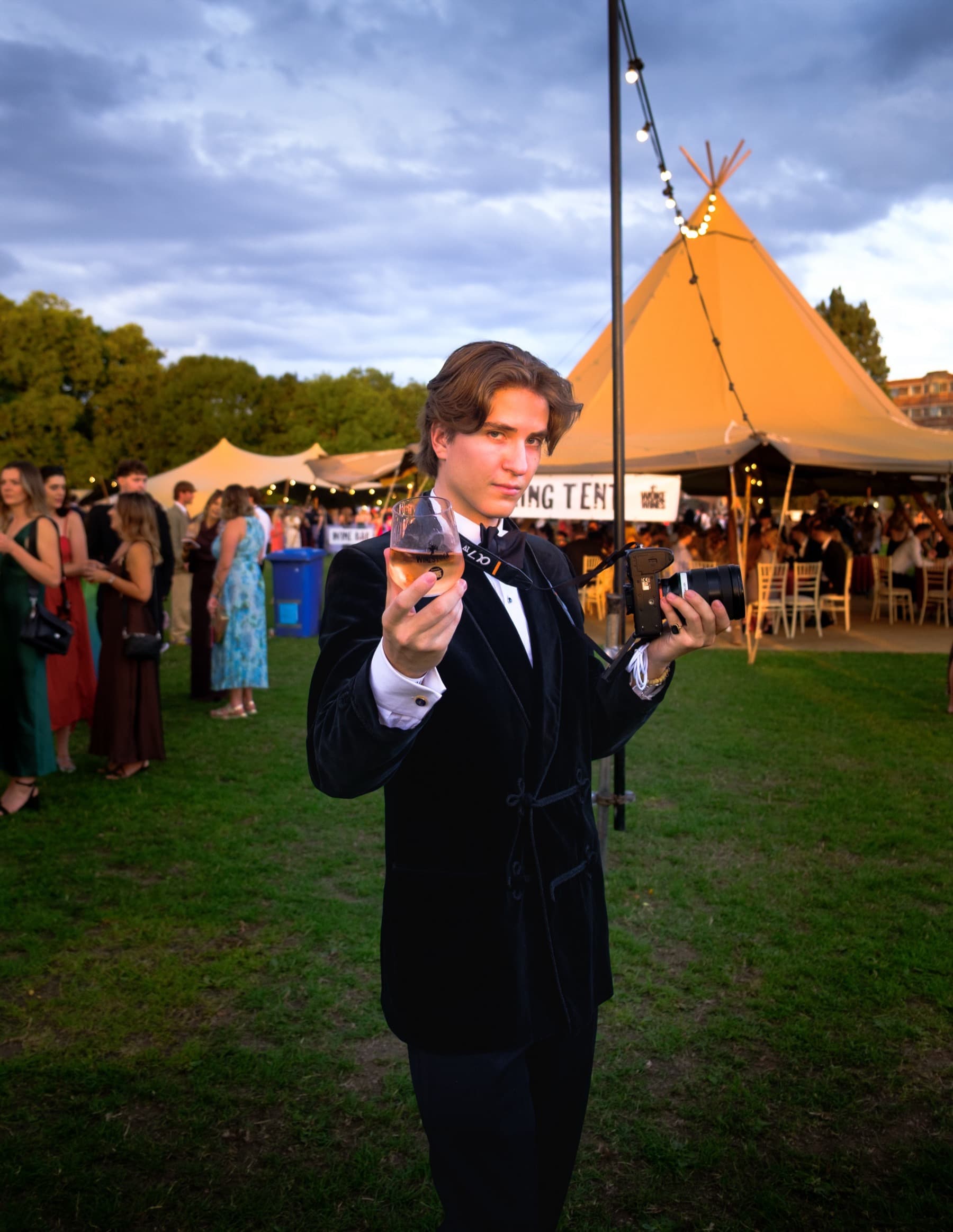 Guest in a velvet blazer holding a glass of wine with a tipi tent and festoon lights behind at dusk