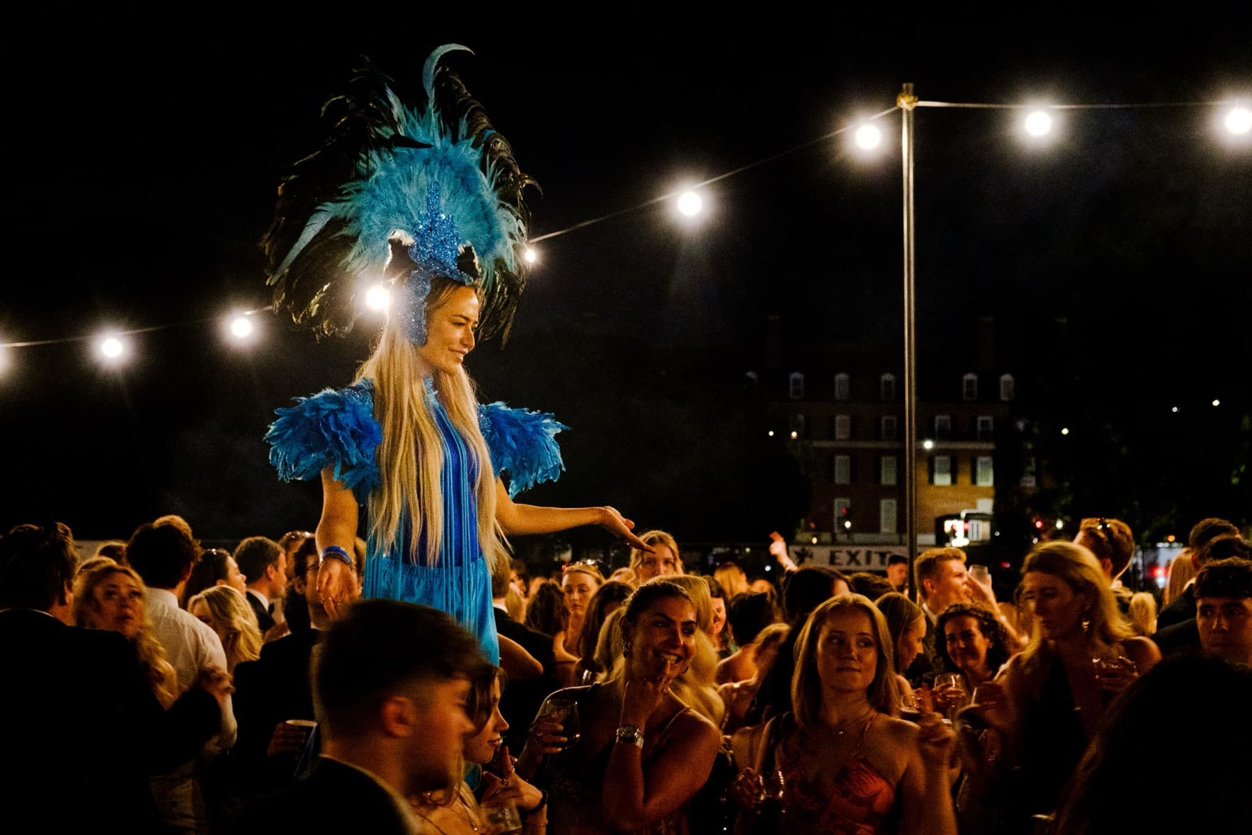 Performer in a blue feathered headdress and costume elevated above the crowd under festoon lights at night