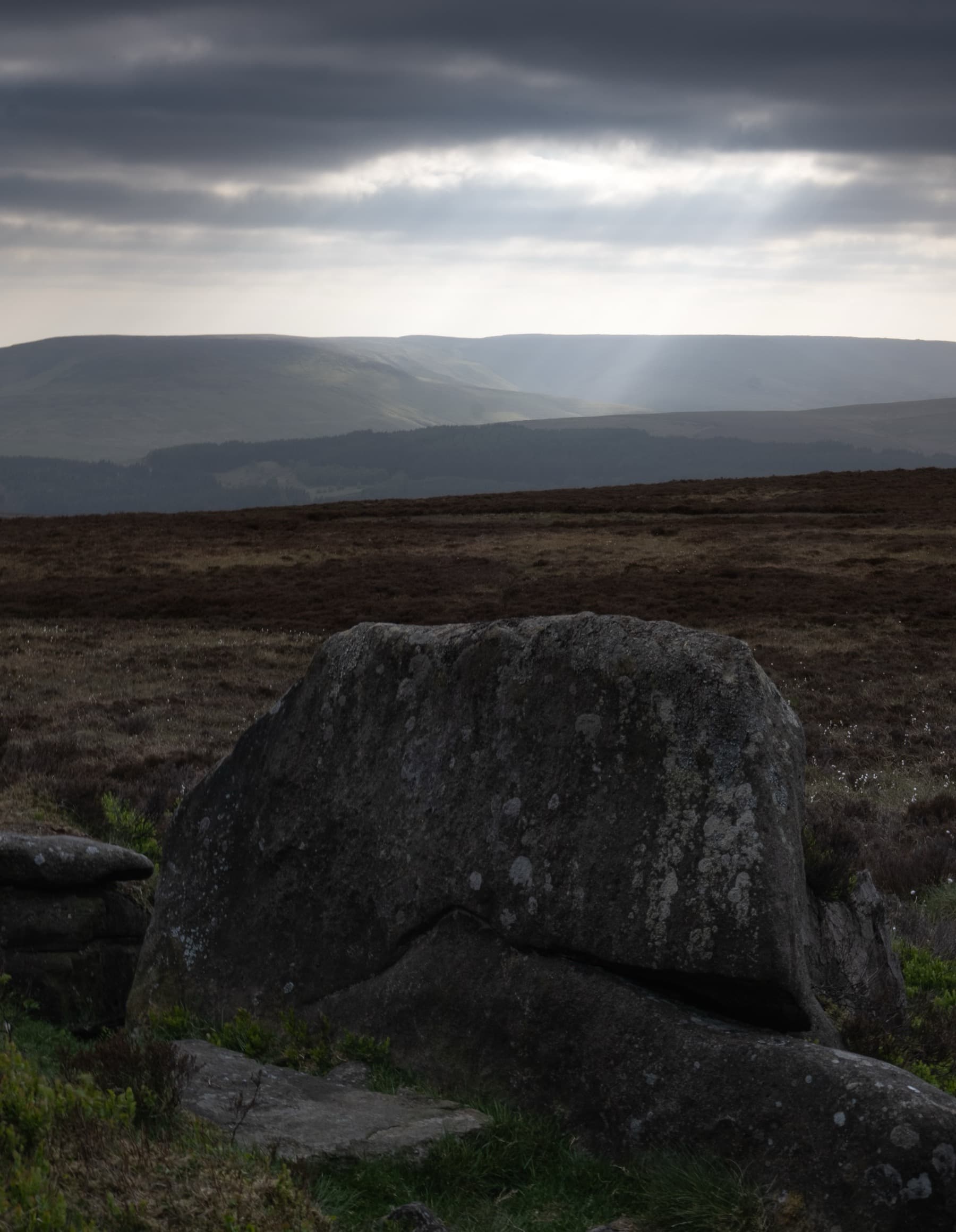 Large gritstone boulder on moorland with sun rays breaking through clouds over the distant hills
