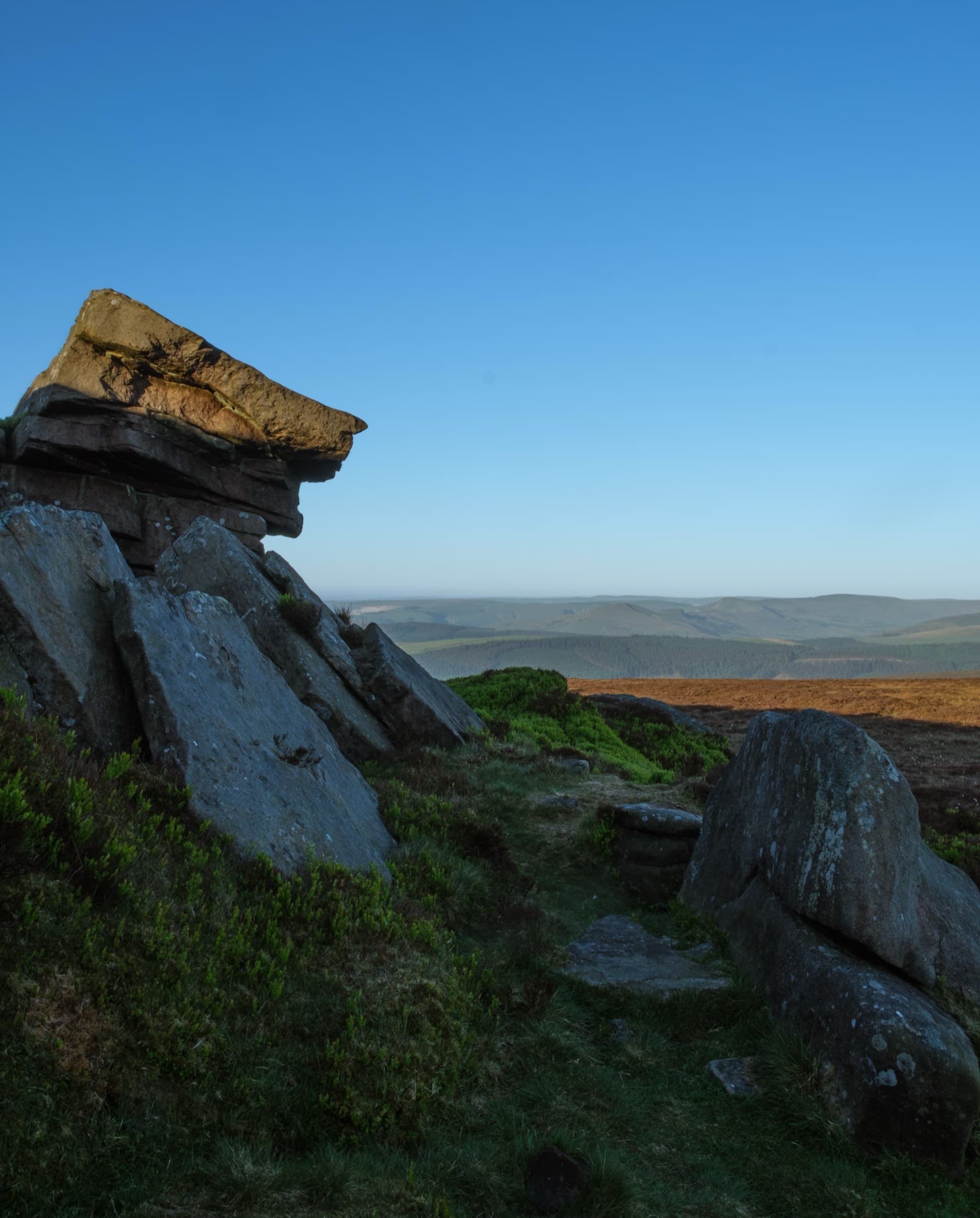 Weathered gritstone outcrop with a sunlit capstone perched on top overlooking rolling Peak District moorland under a blue sky