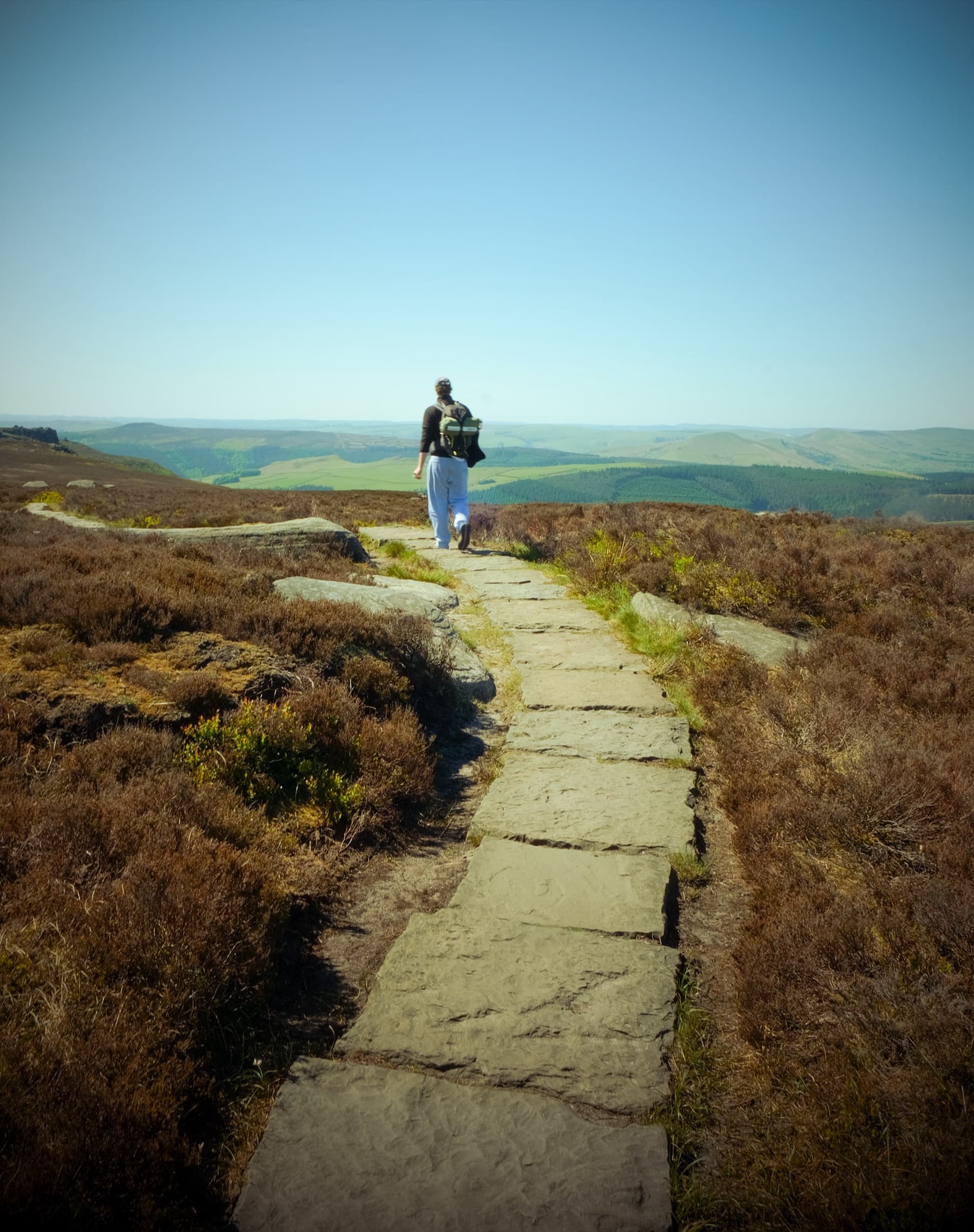 Hiker walking along a flagstone path through heather moorland toward distant Peak District hills under a clear sky
