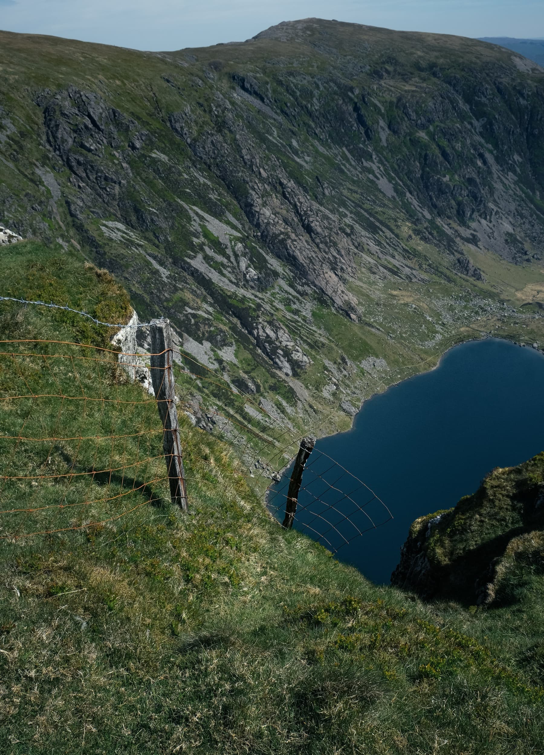 Fence post and wire on a grassy ridge with a steep drop to a dark mountain lake below and cliffs beyond