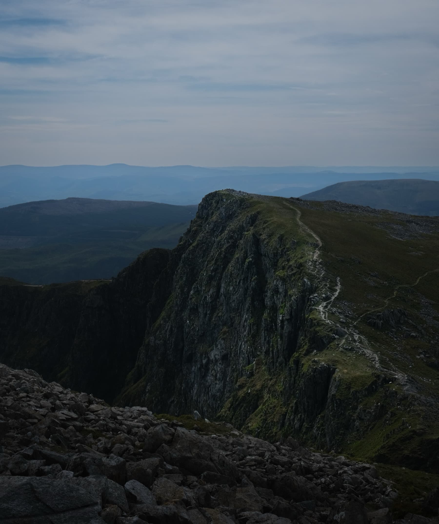 Steep cliff edge with a trail along the summit ridge looking out over hazy hills under a clear sky