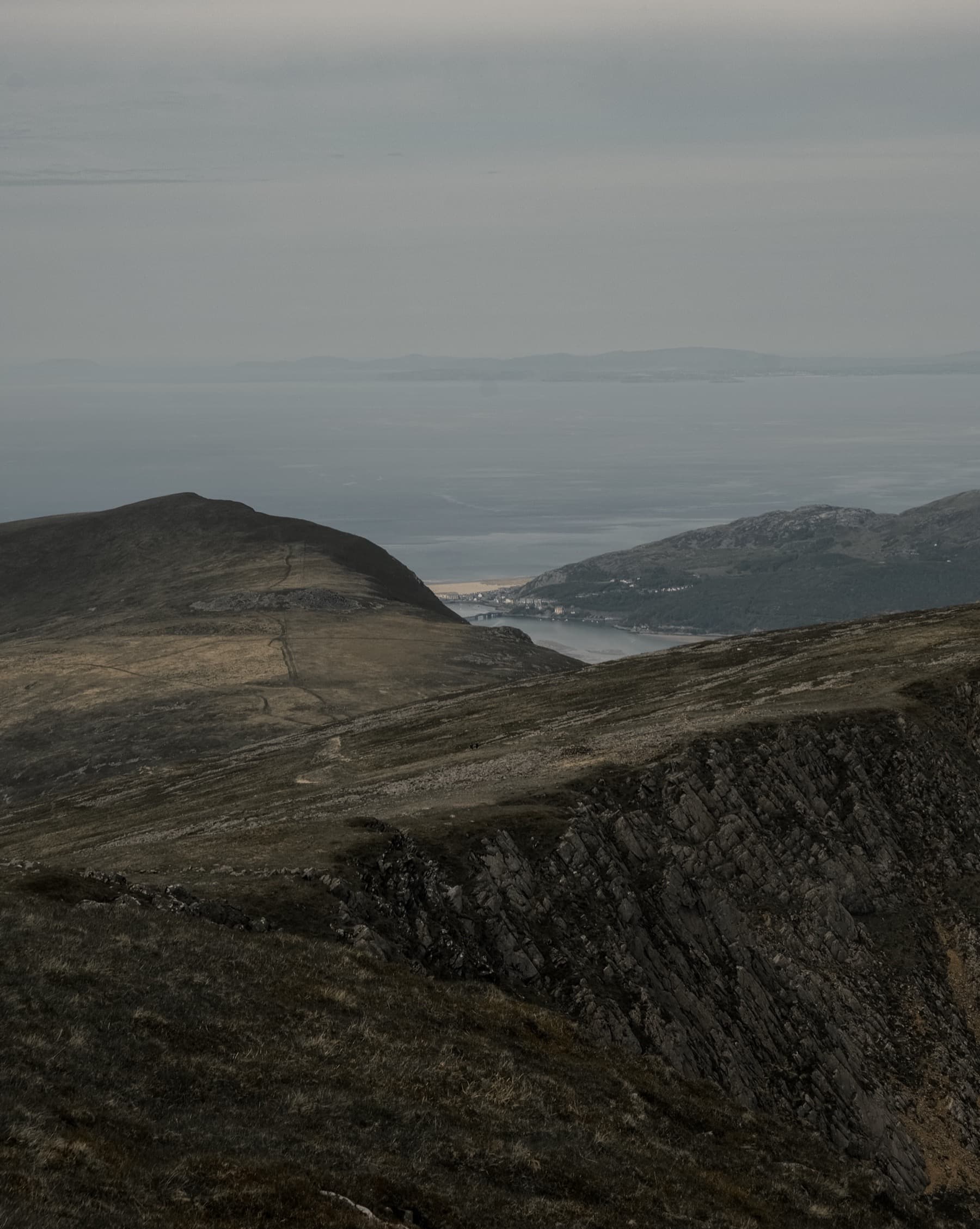 View from the mountain summit over rolling hills toward a distant estuary and coastline in hazy light