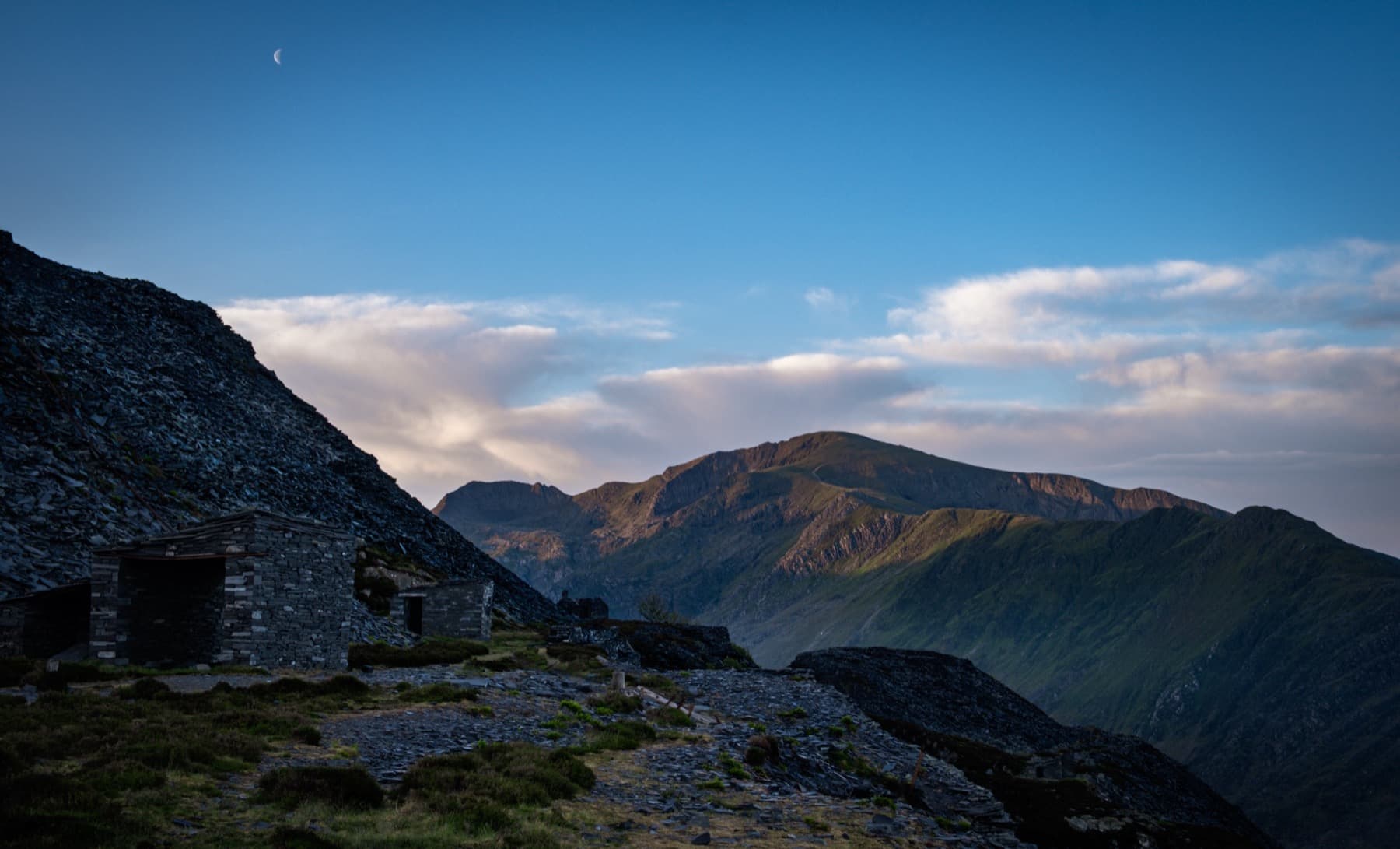 Ruined slate quarry buildings at twilight with a half moon above and Snowdon range lit by the last golden light