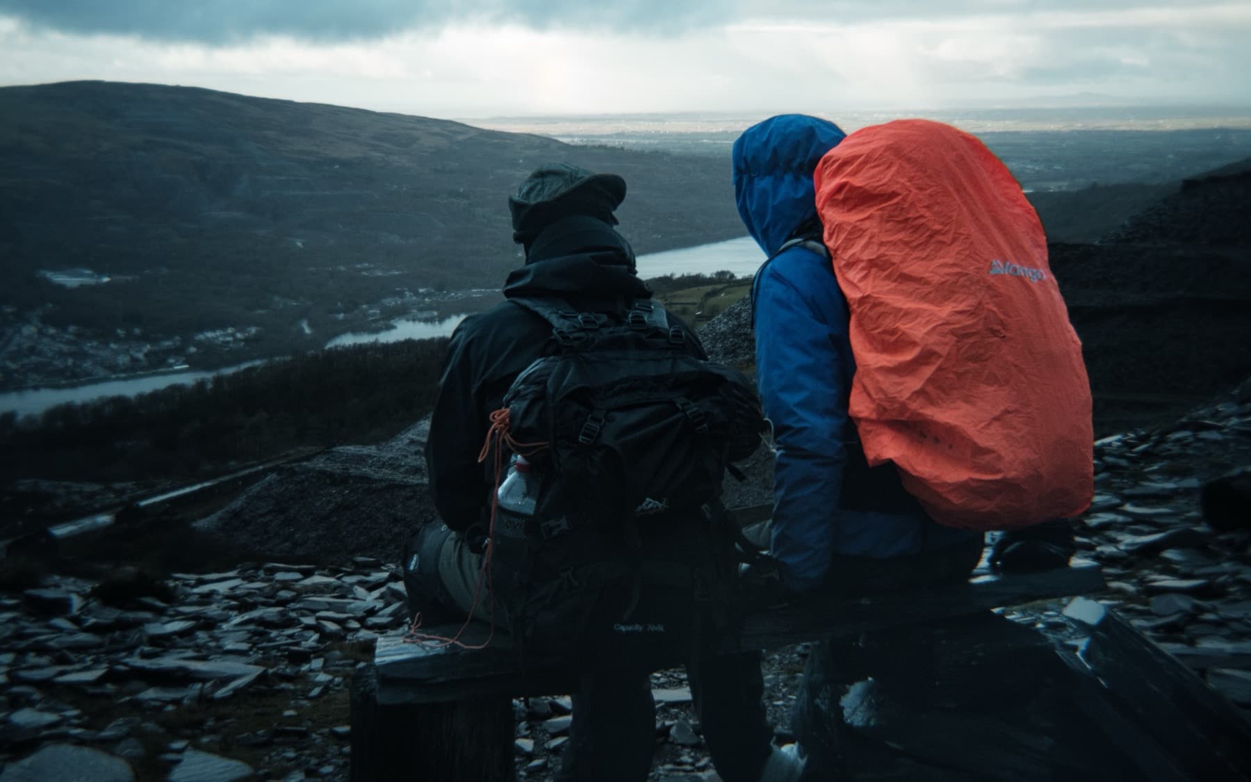 Two hikers with large backpacks sitting on slate and looking out over a valley and lake from a quarry ledge