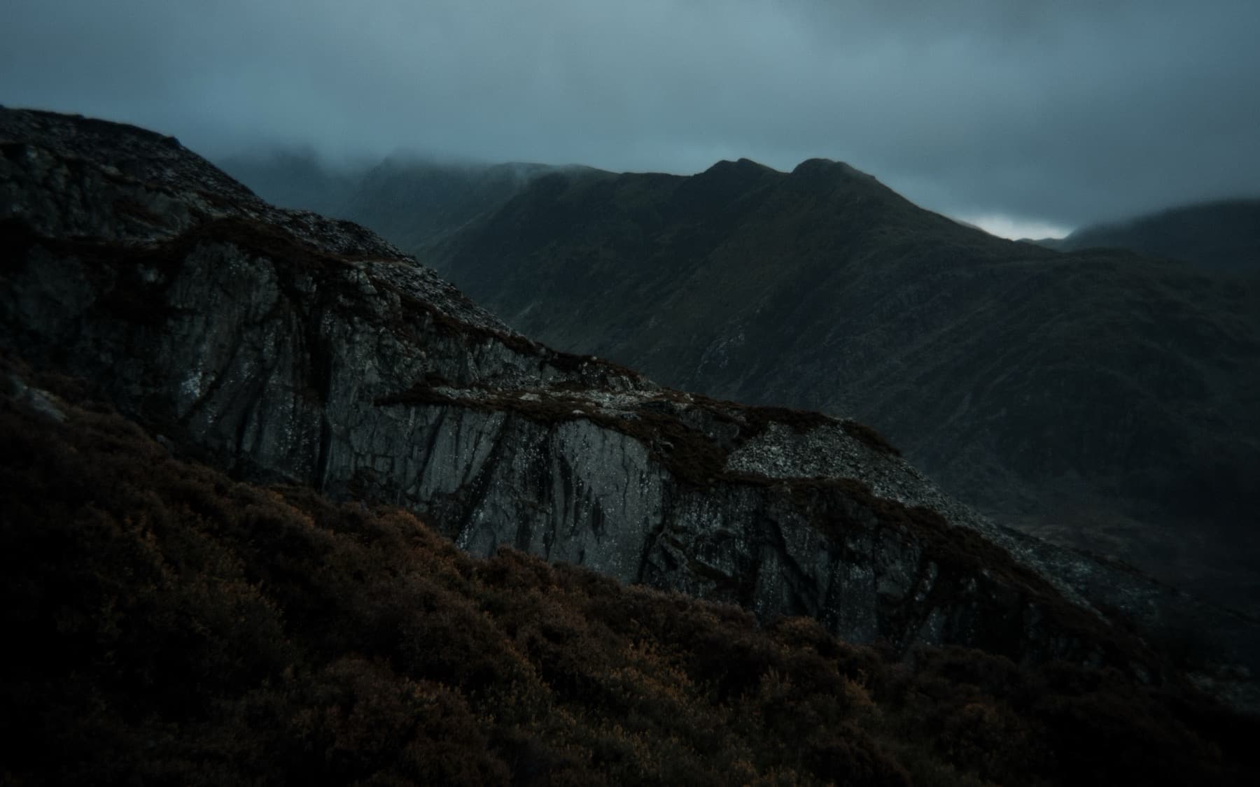 Dark slate rock face with heather in the foreground and cloud-shrouded mountains behind under an overcast sky