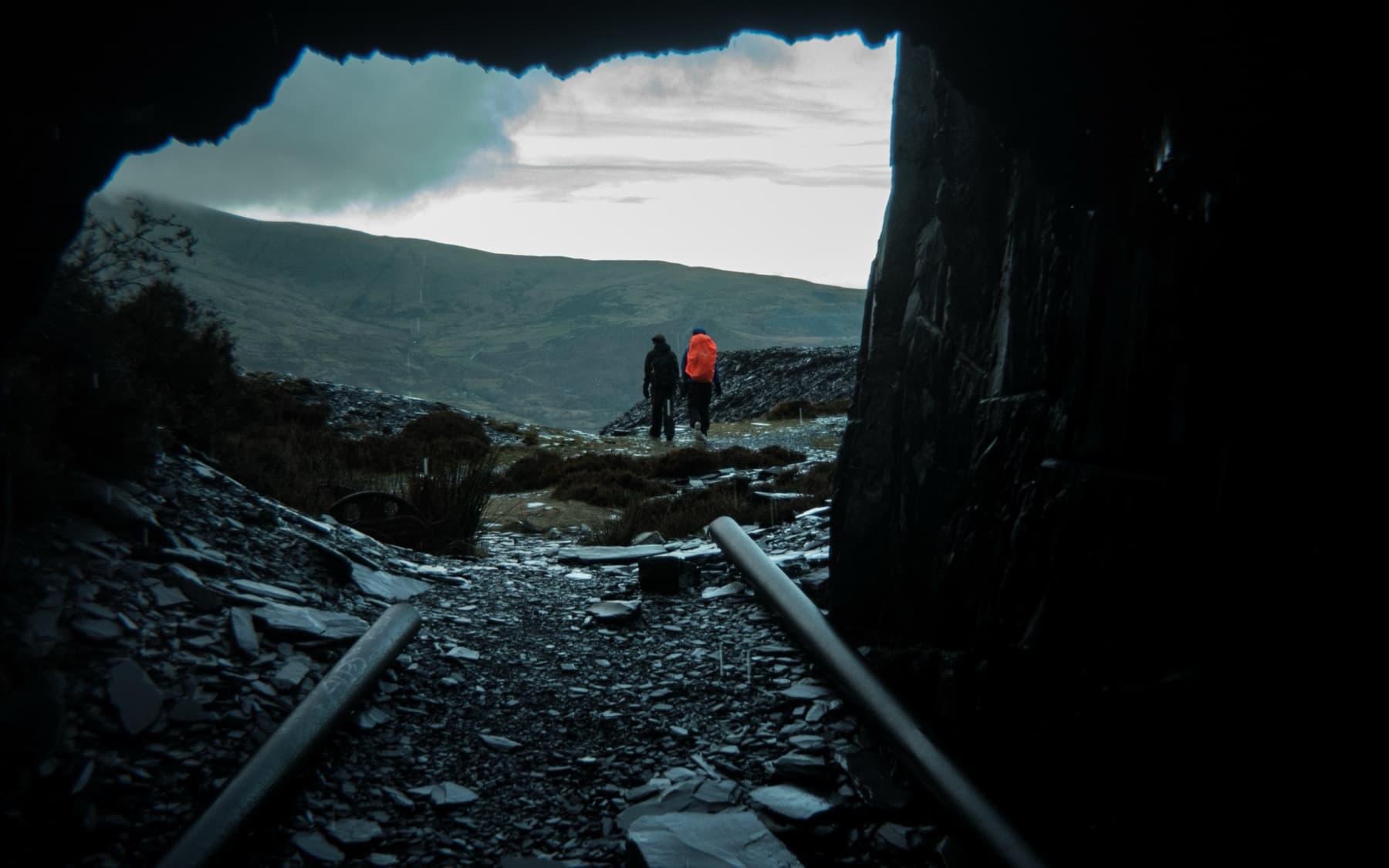 Two hikers emerging from the dark mouth of a quarry tunnel along old rail tracks into overcast mountain landscape