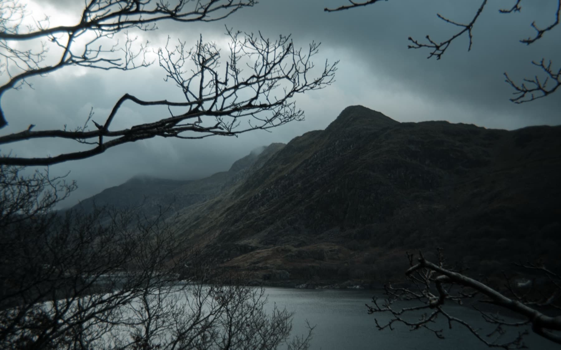 Mountain and lake framed through bare winter tree branches under dramatic dark clouds
