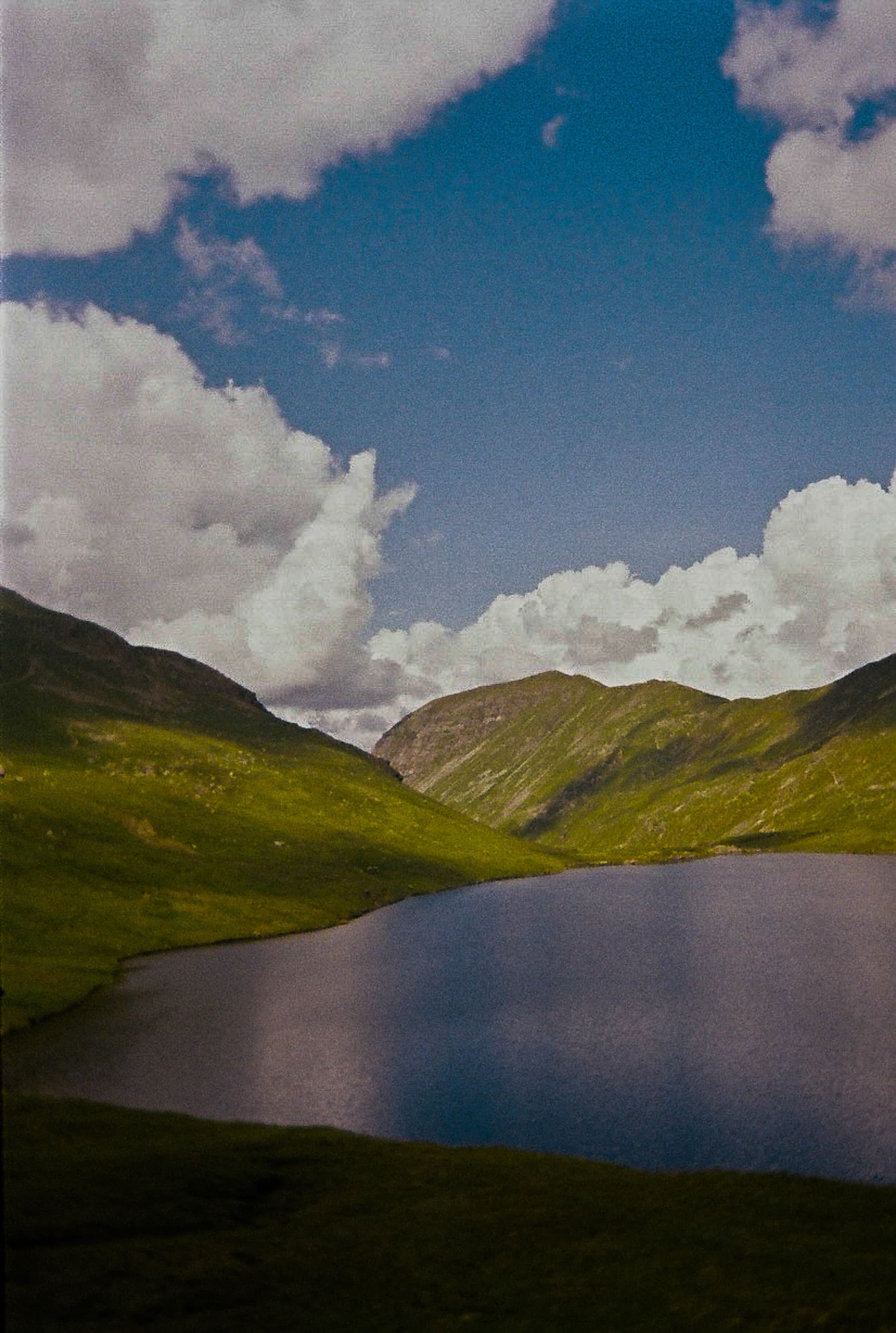 Mountain tarn nestled between green hillsides reflecting white cumulus clouds under a blue sky, shot on film