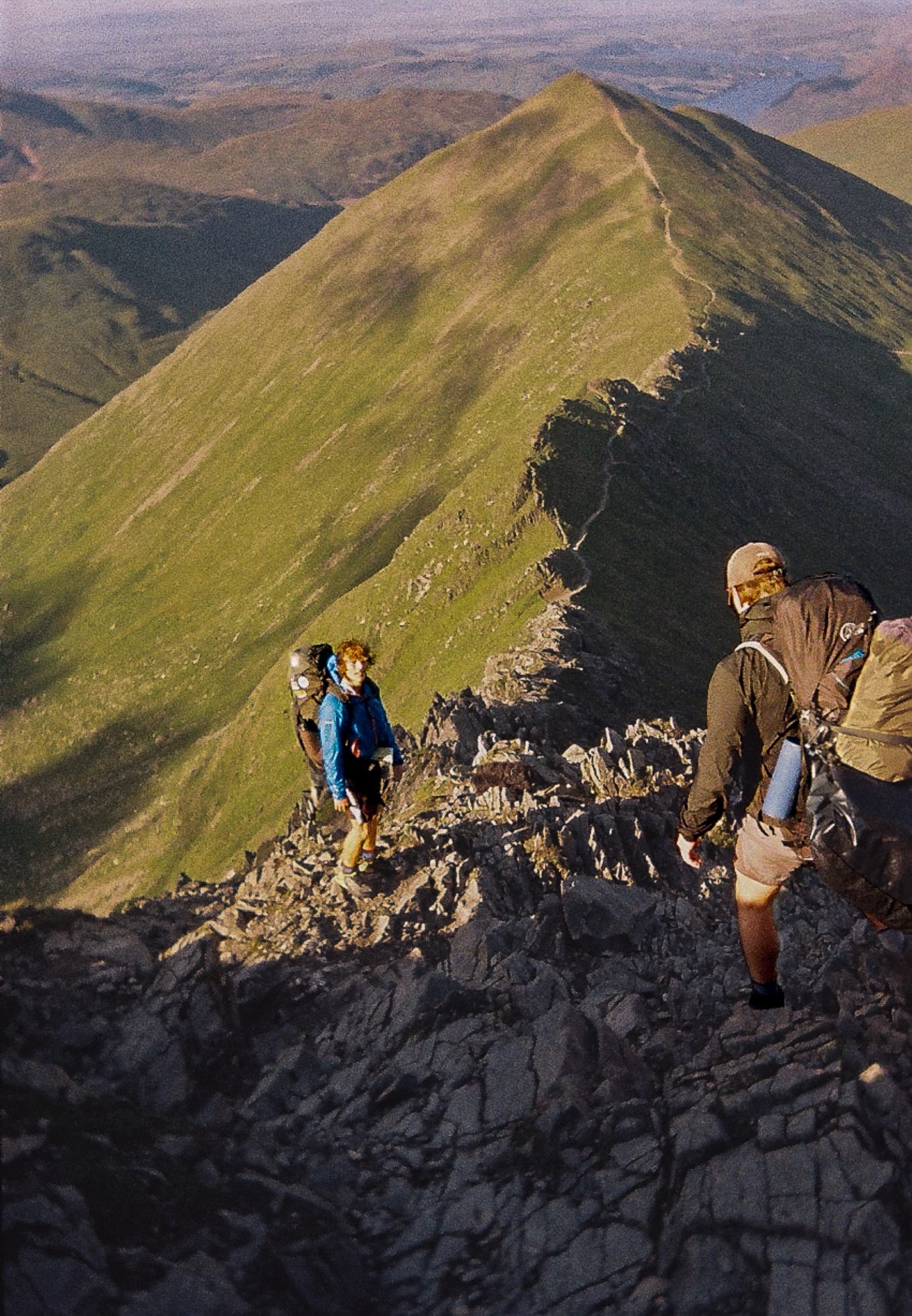 Two hikers scrambling along a narrow rocky ridge with steep green slopes falling away on both sides, shot on film