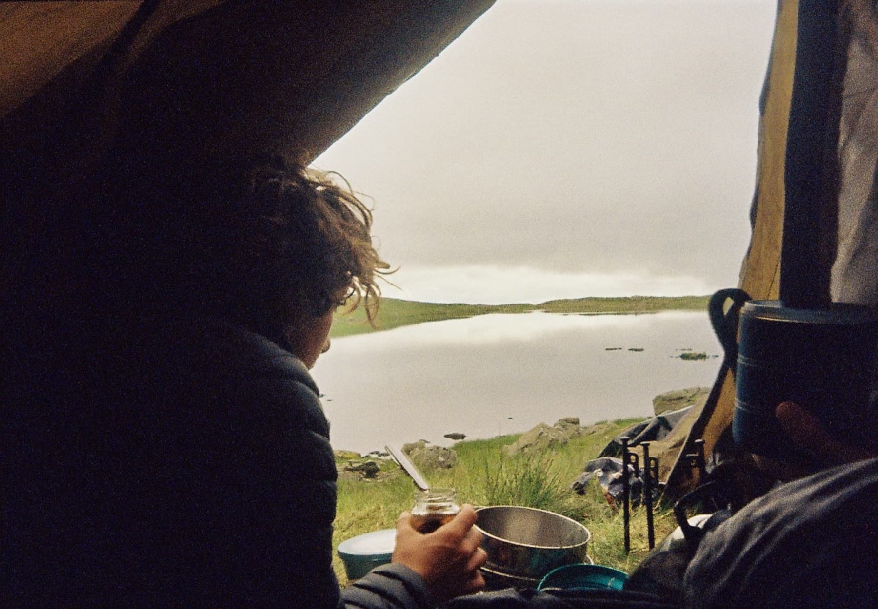 Person looking out from a tent at a calm mountain lake while holding a mug with camping gear nearby, shot on film