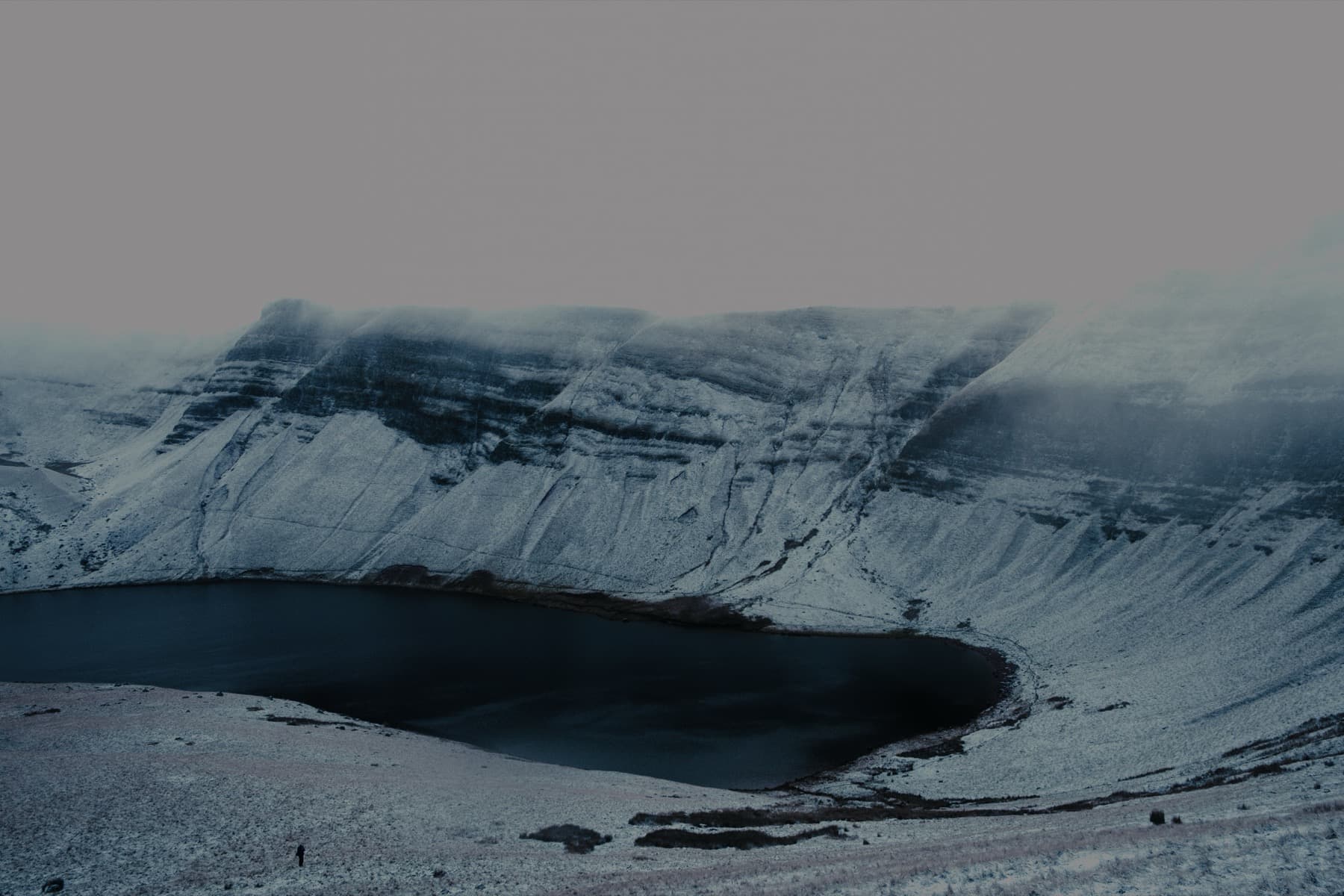 Snow-covered escarpment curving around the dark waters of Llyn y Fan Fach with mist rolling over the ridge