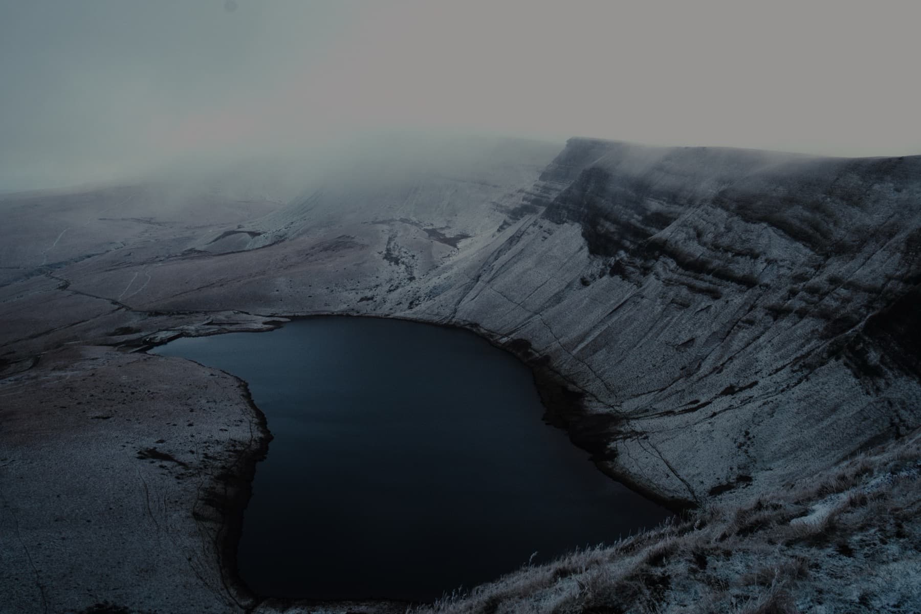Aerial view of dark glacial lake nestled beneath frost-covered cliffs disappearing into low cloud