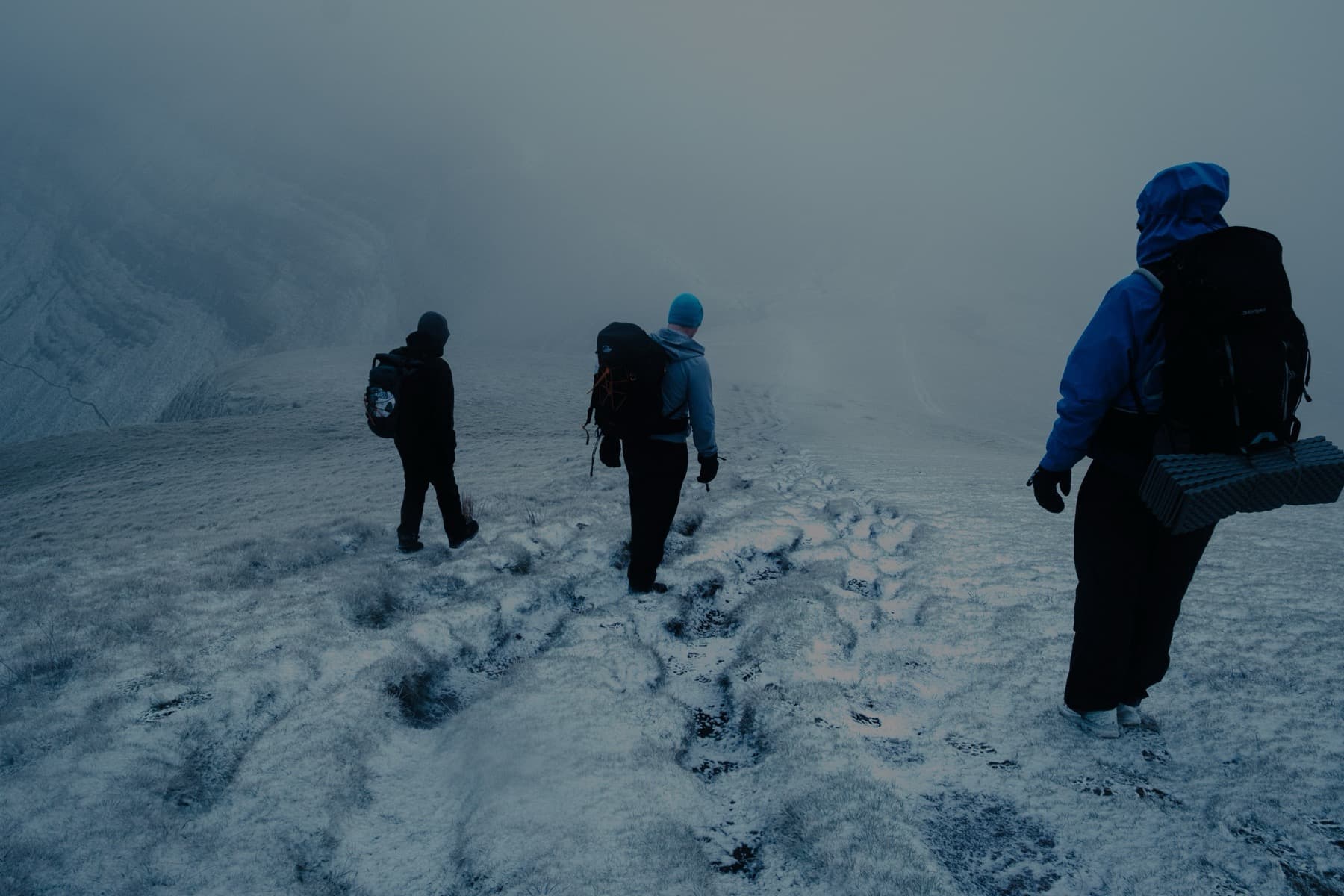 Three hikers descending a snowy slope into thick fog with a mountain escarpment barely visible behind