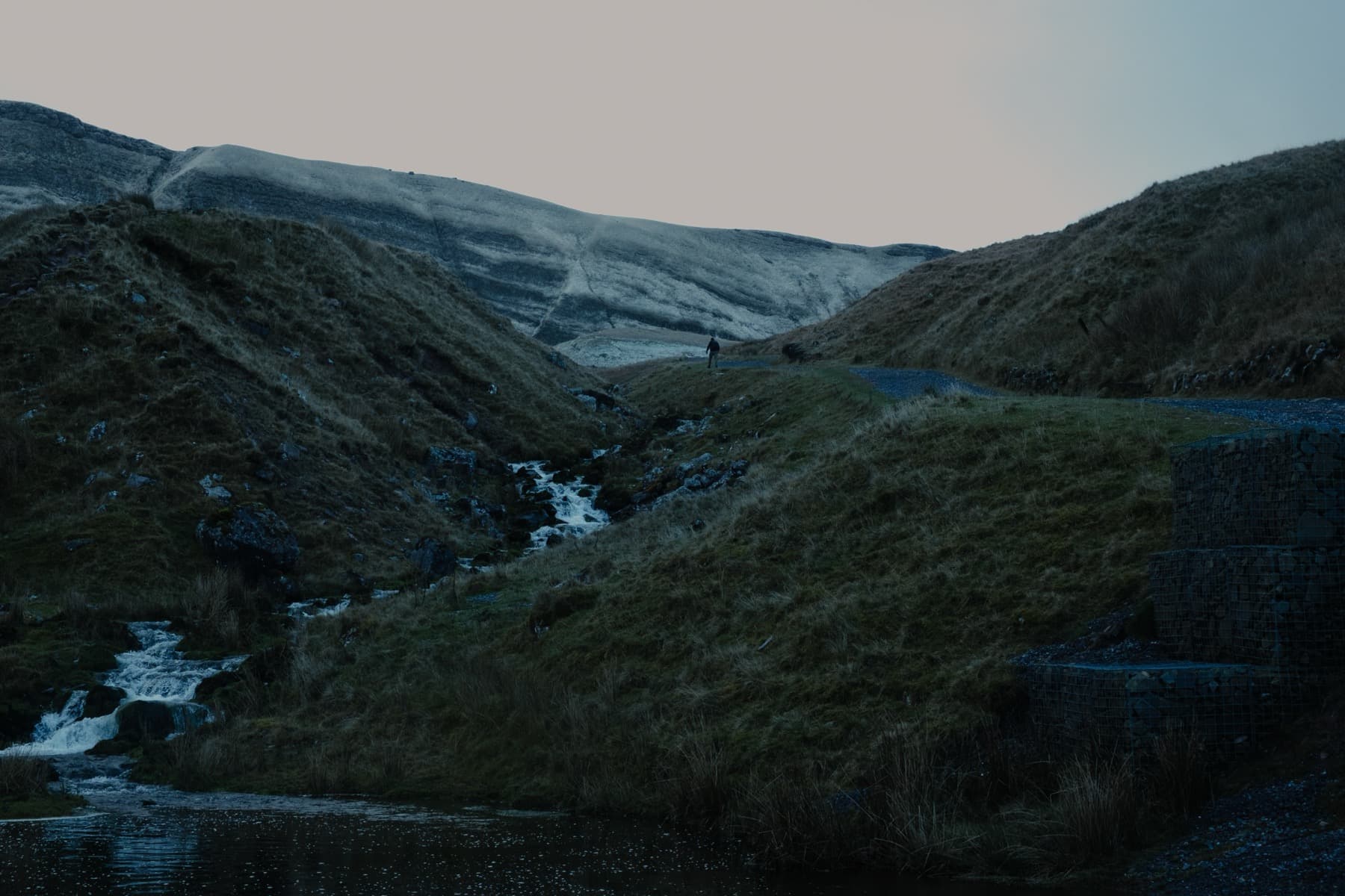 Lone figure walking along a road in a grassy valley with a stream and snow-capped mountains in the fading light
