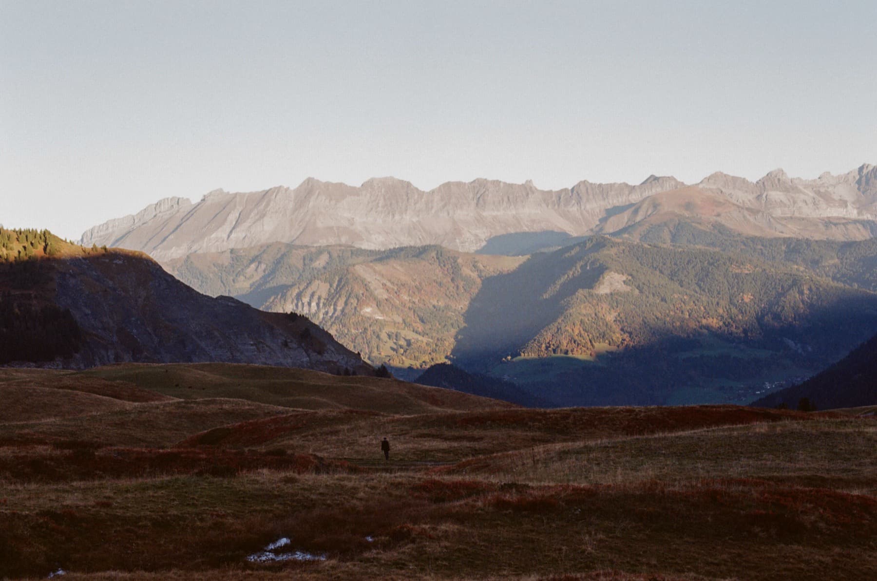 A lone figure standing on a vast autumn alpine meadow with a dramatic wall of limestone cliffs and layered valleys stretching into the distance, shot on film