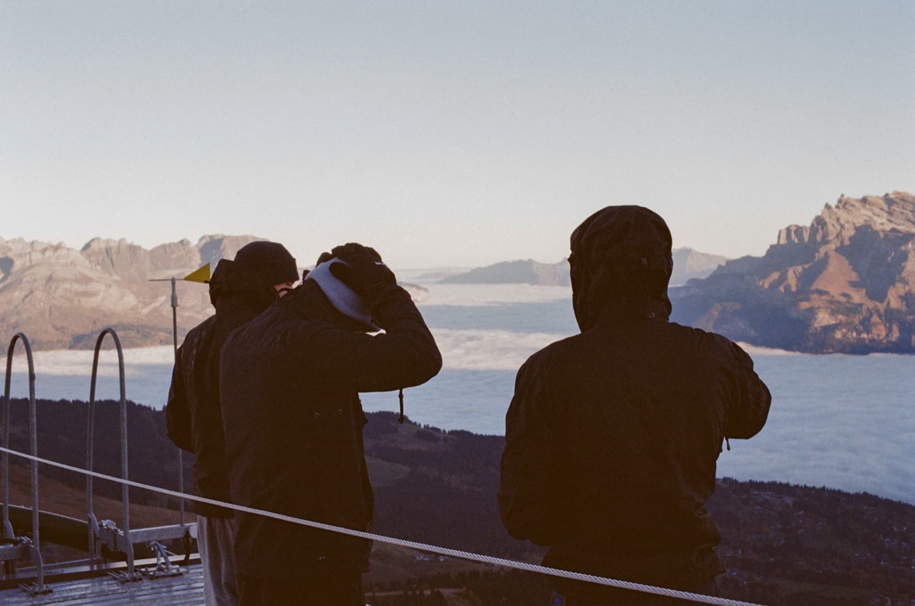 Two silhouetted figures photographing a cloud inversion and distant peaks from a metal viewing platform at dawn, shot on film