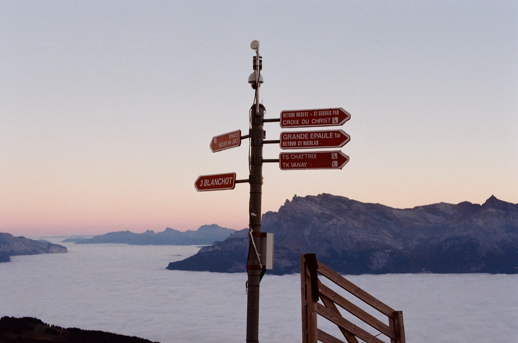 Hiking trail signpost above a cloud inversion at sunset with signs for Megeve, Mont Joly, Grande Epaule, and Blanchot, mountain peaks in the pink twilight beyond, shot on film