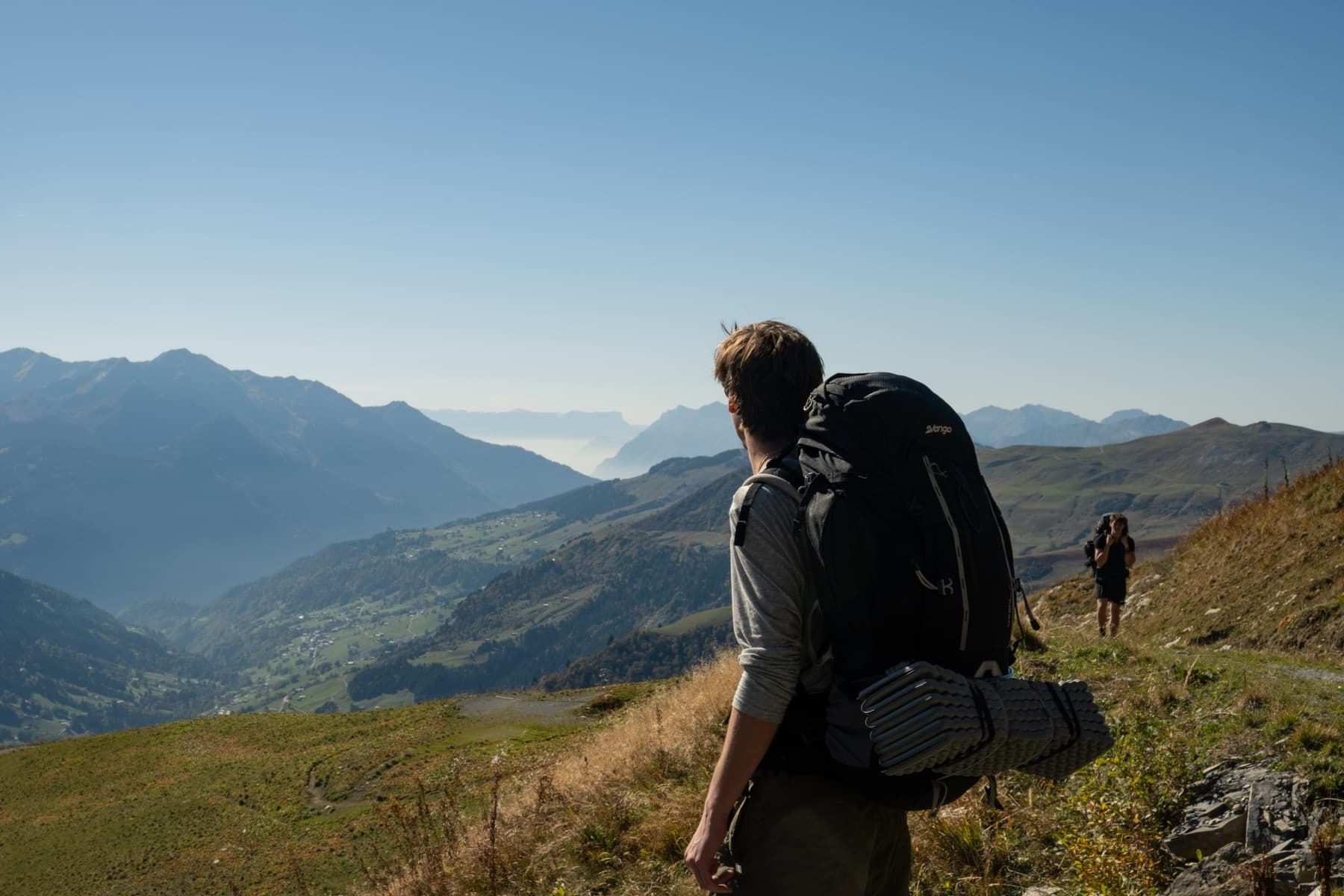 A hiker with a large backpack and sleeping mat looking out over a sweeping valley and layered hazy mountains on the Mont Joly trail