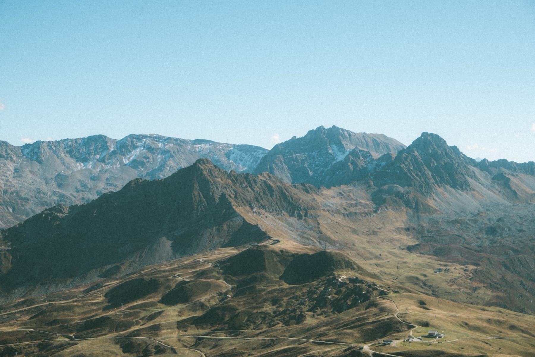Sweeping panoramic view from a high alpine ridge showing barren mountain slopes, winding paths, and distant snow-dusted peaks under a clear blue sky