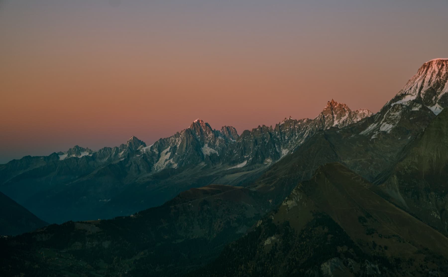 Jagged Alpine peaks and the snow-capped Mont Blanc massif silhouetted against a warm orange and pink sunset sky