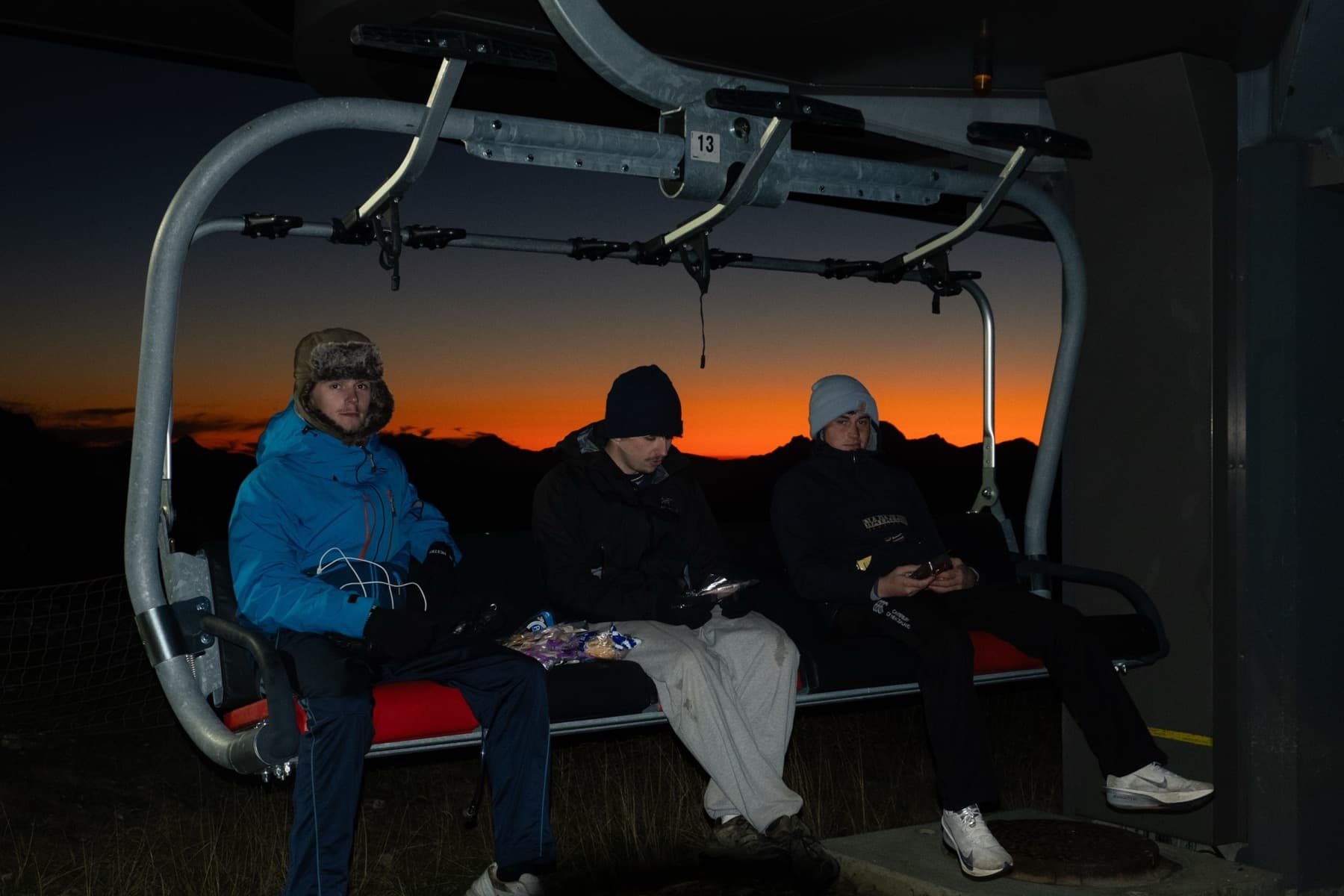 Three friends bundled in warm jackets sitting on a stationary chairlift seat at dusk with a vivid orange sunset behind the mountains