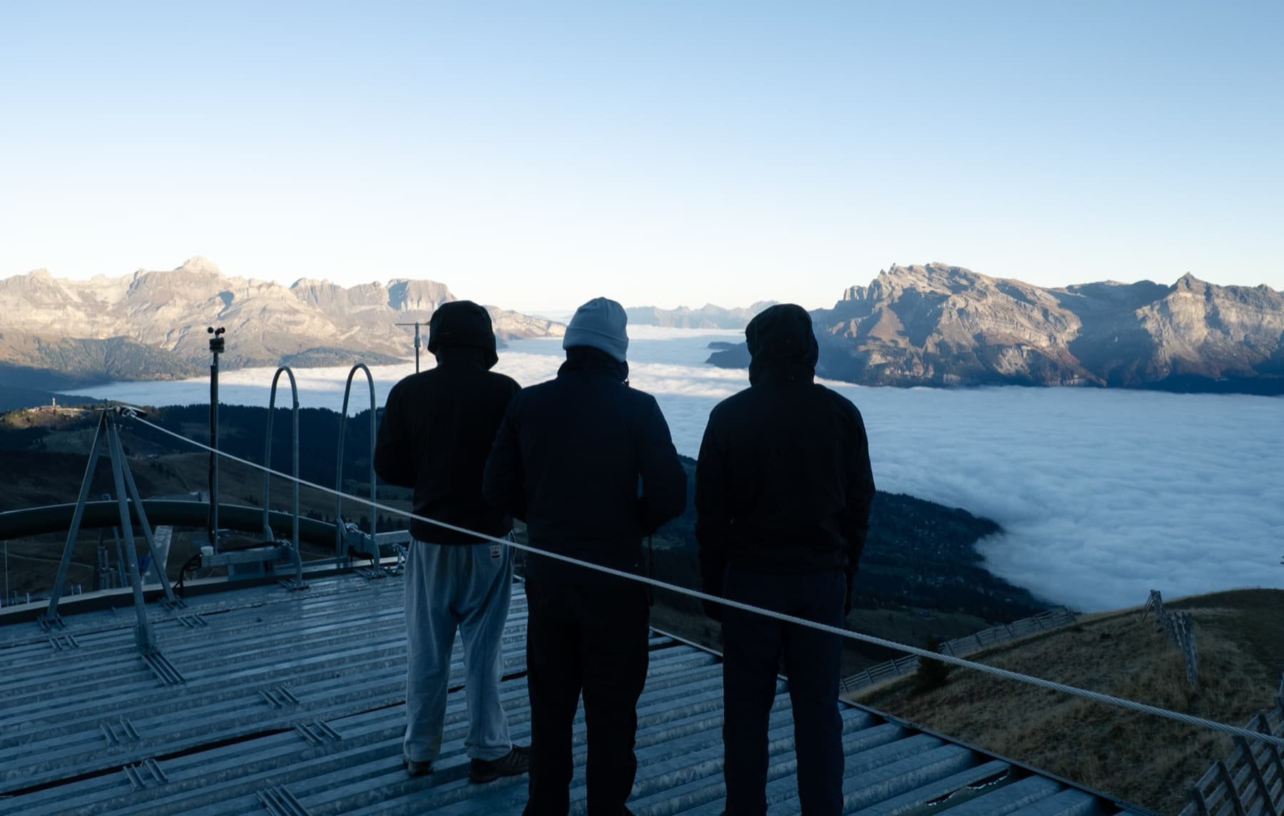 Three silhouetted figures standing on a metal platform overlooking a thick cloud inversion filling the valley between sunlit Alpine peaks at dawn