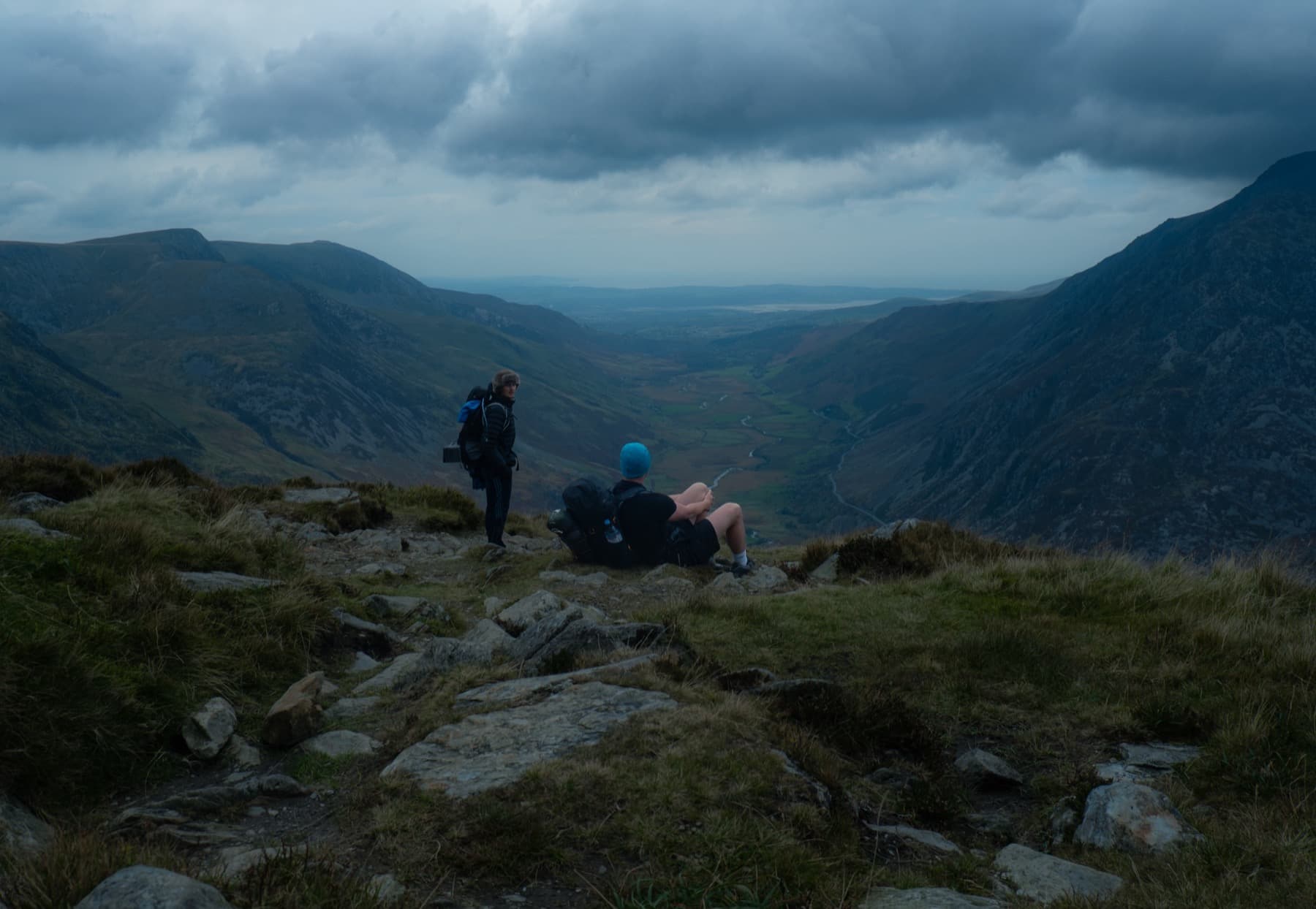 Two hikers resting on a rocky summit overlooking a wide U-shaped valley stretching toward the coast under heavy cloud