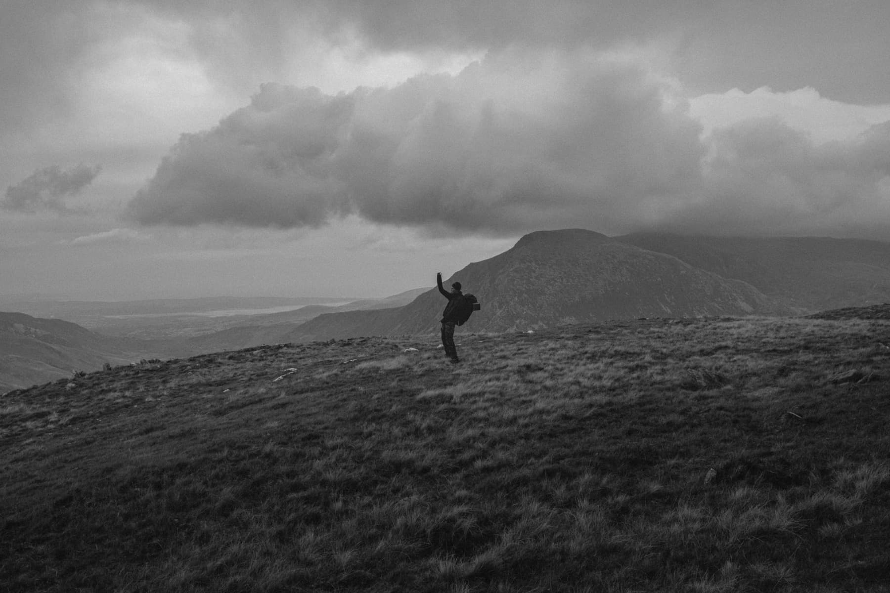 Hiker leaning into the wind on an exposed grassy ridge in black and white with arm raised against dramatic sky