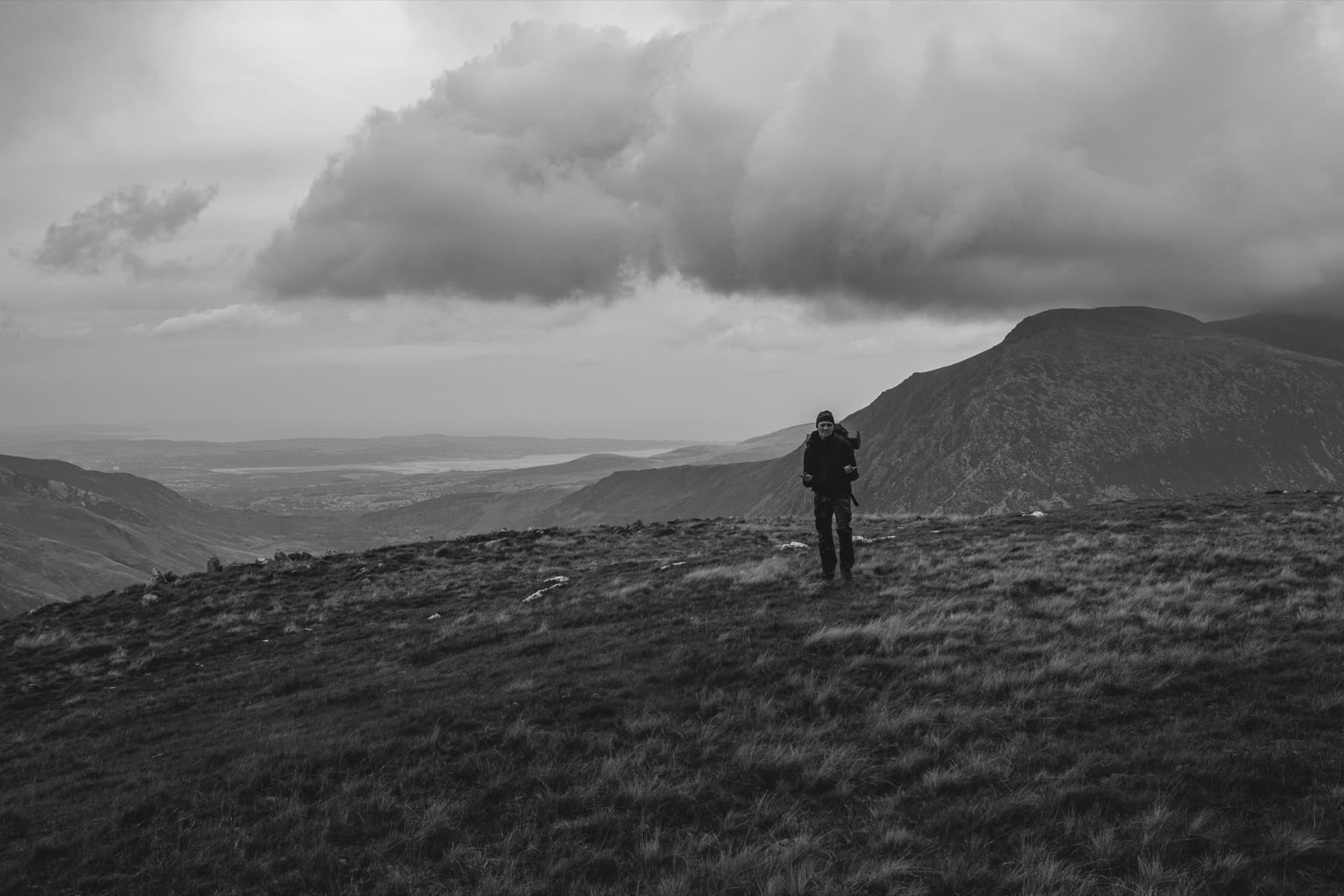 Hiker with backpack walking across open moorland in black and white with mountains and dramatic clouds behind