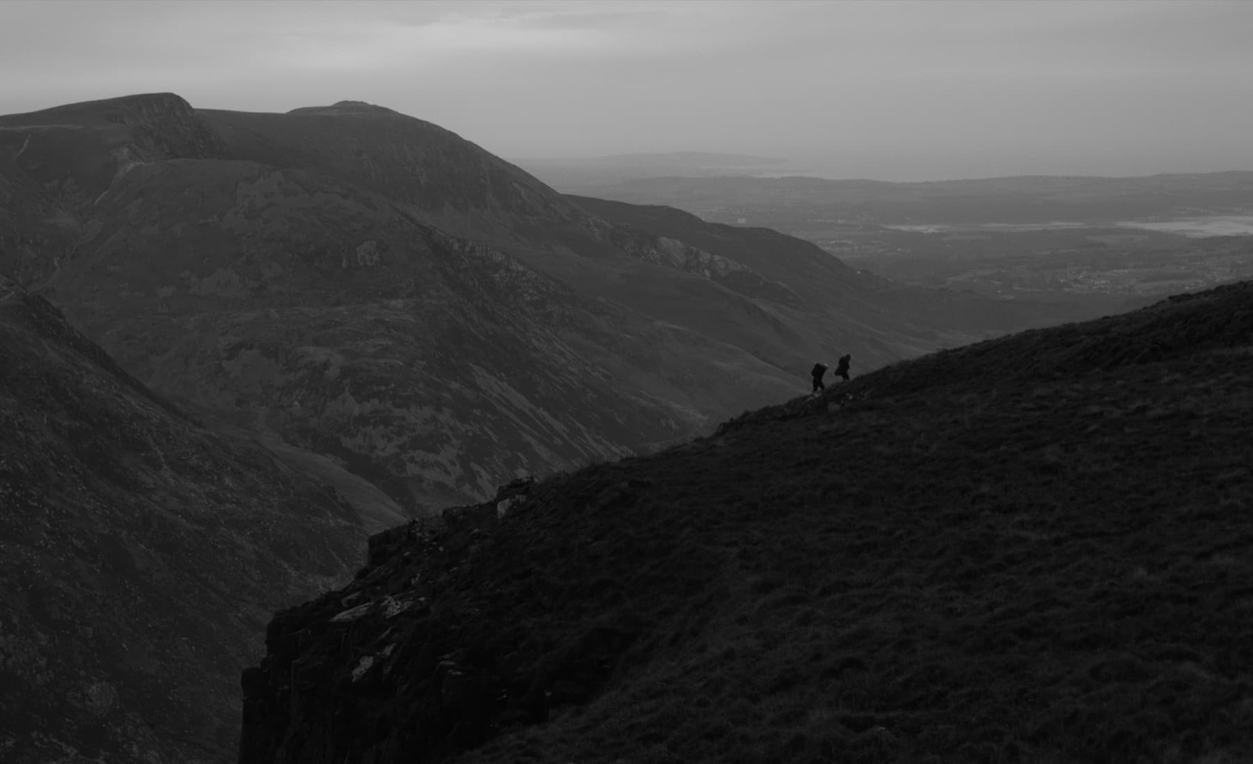 Two silhouetted hikers ascending a steep grassy ridge with mountains stretching into the distance in black and white