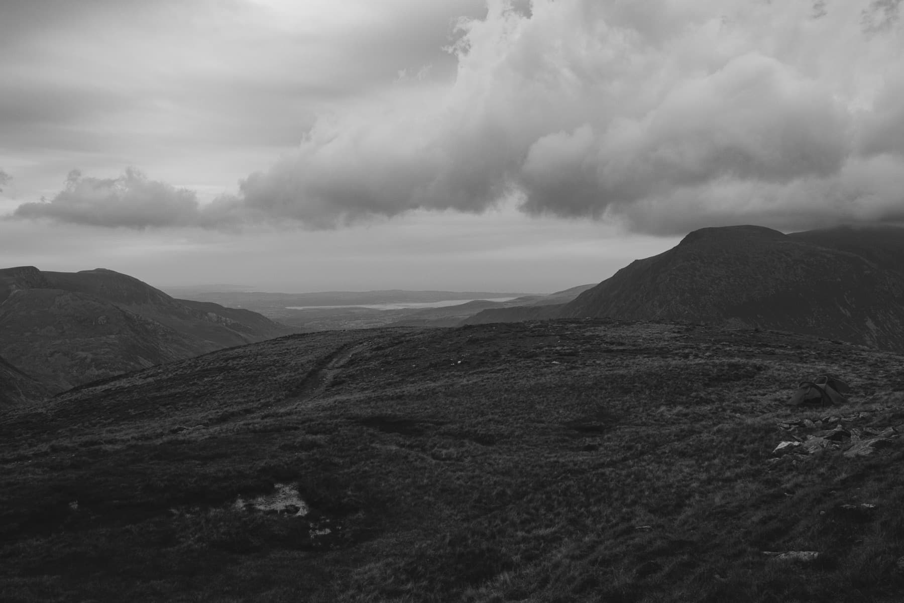 Wide moorland plateau between mountains with a wild camp tent visible and dramatic clouds gathering overhead in black and white